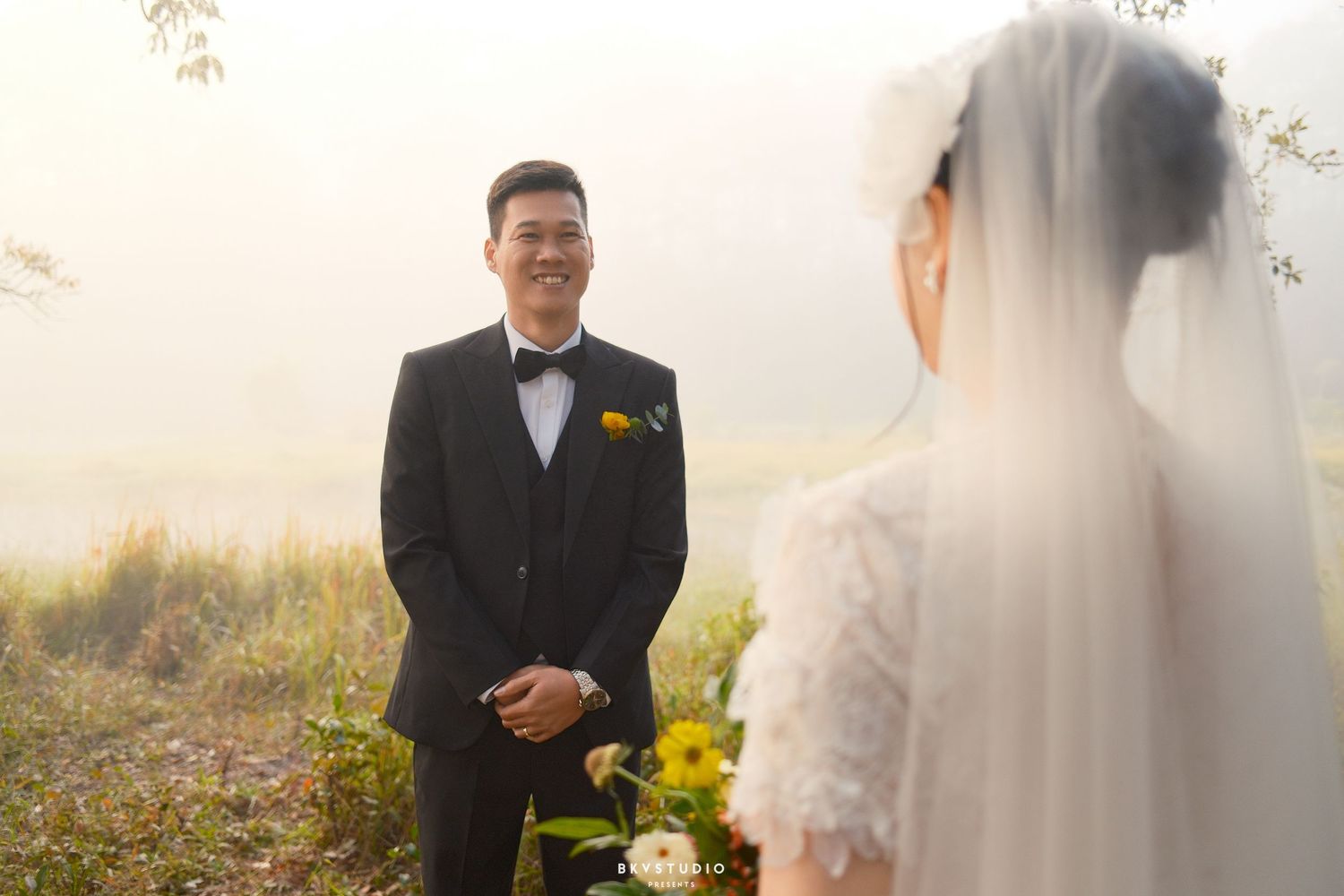Groom in black tuxedo smiles at bride in white lace wedding dress and veil during outdoor ceremony in misty field.