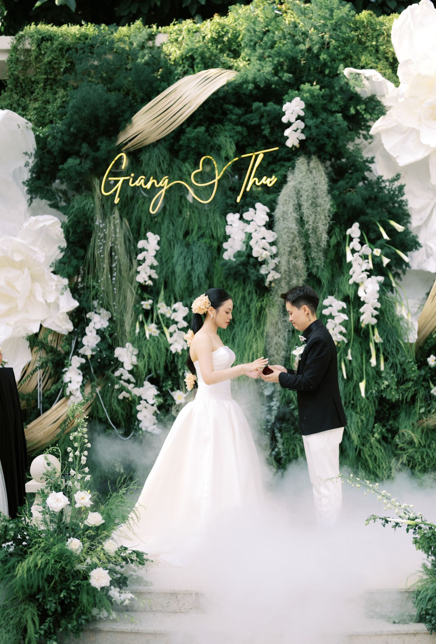 Couple exchanging rings at outdoor garden wedding ceremony under floral arch with Giang and Thu neon sign.
