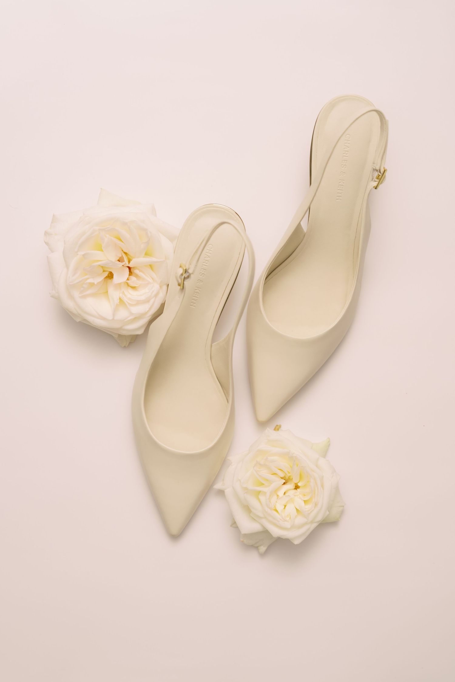 Ivory slingback kitten heels with pointed toes displayed beside white garden roses on a cream background.
