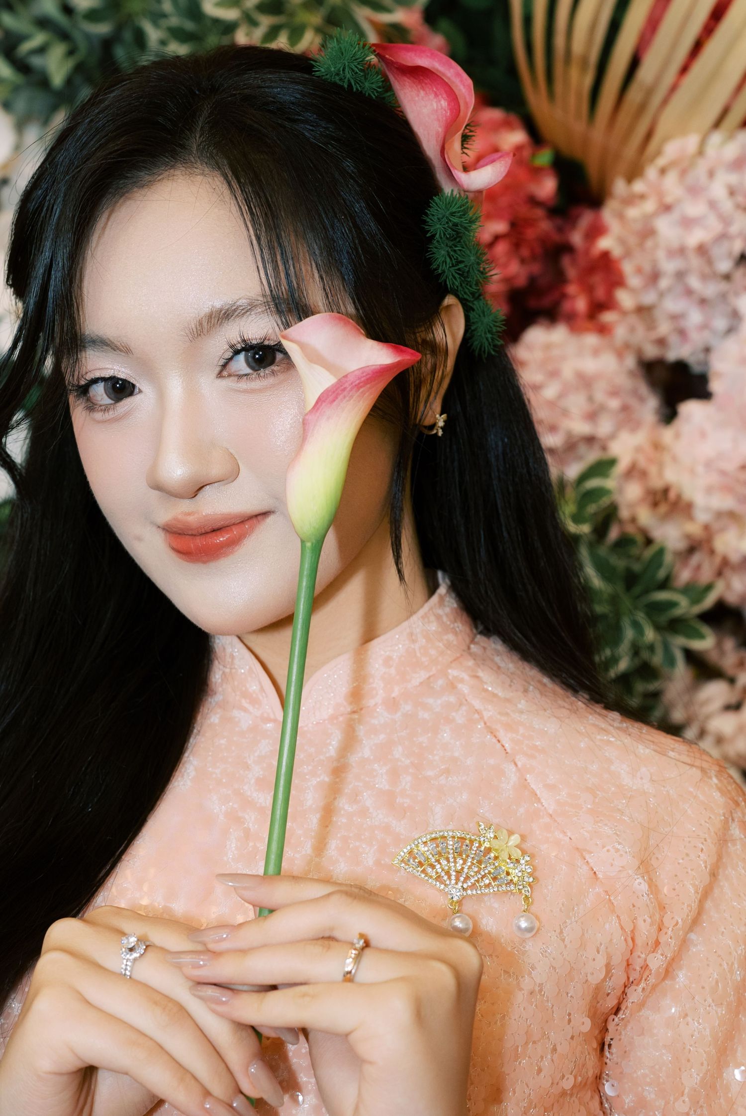 Elegant young woman in pink lace dress holding calla lily with floral hair accessories.