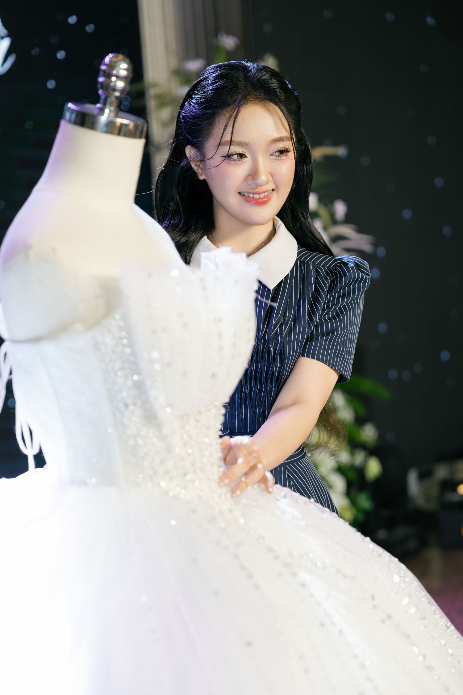Young woman in striped dress smiling while touching a beaded white wedding gown on a mannequin.