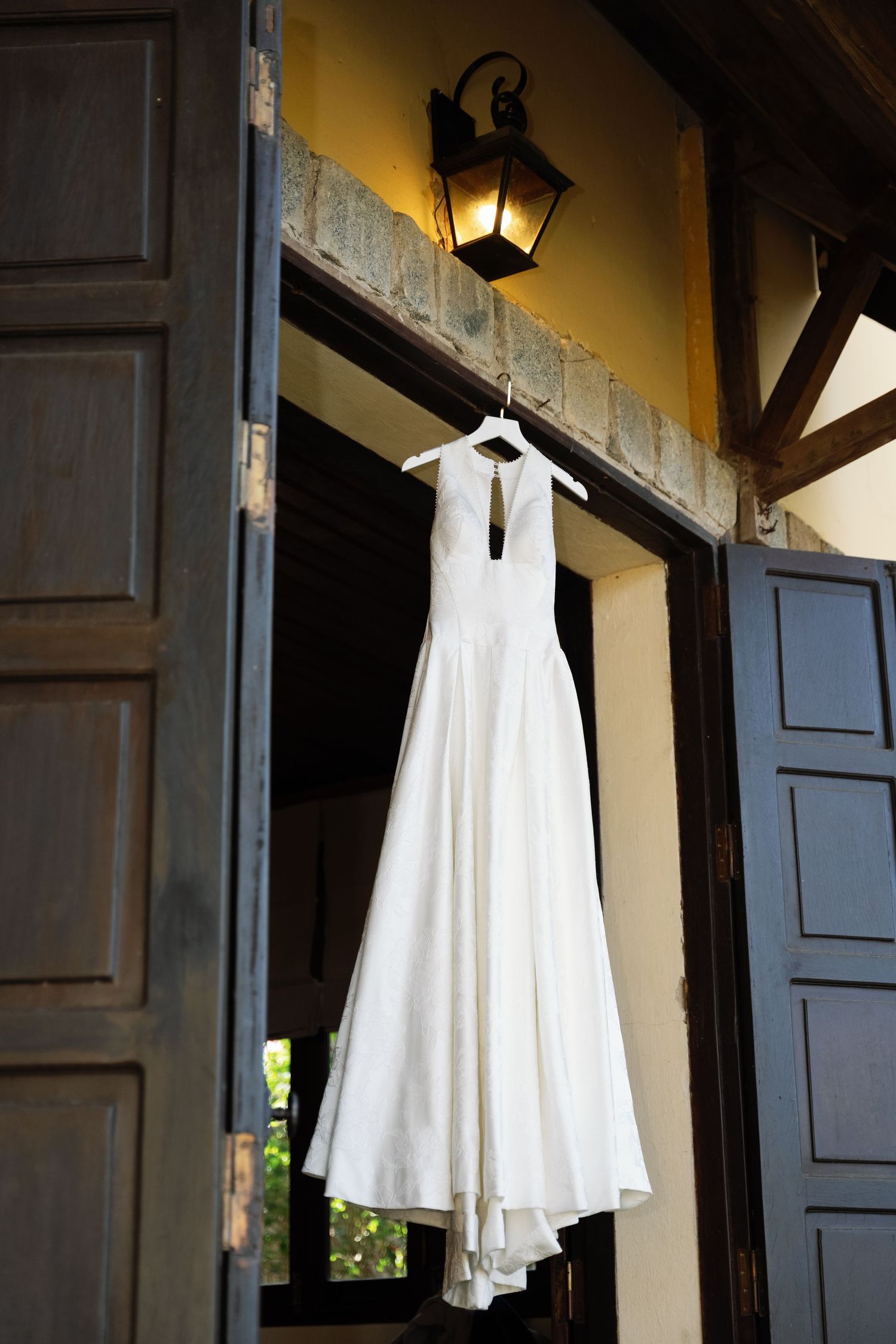 White halter-neck wedding dress hanging on a hanger above rustic wooden doors with lantern.
