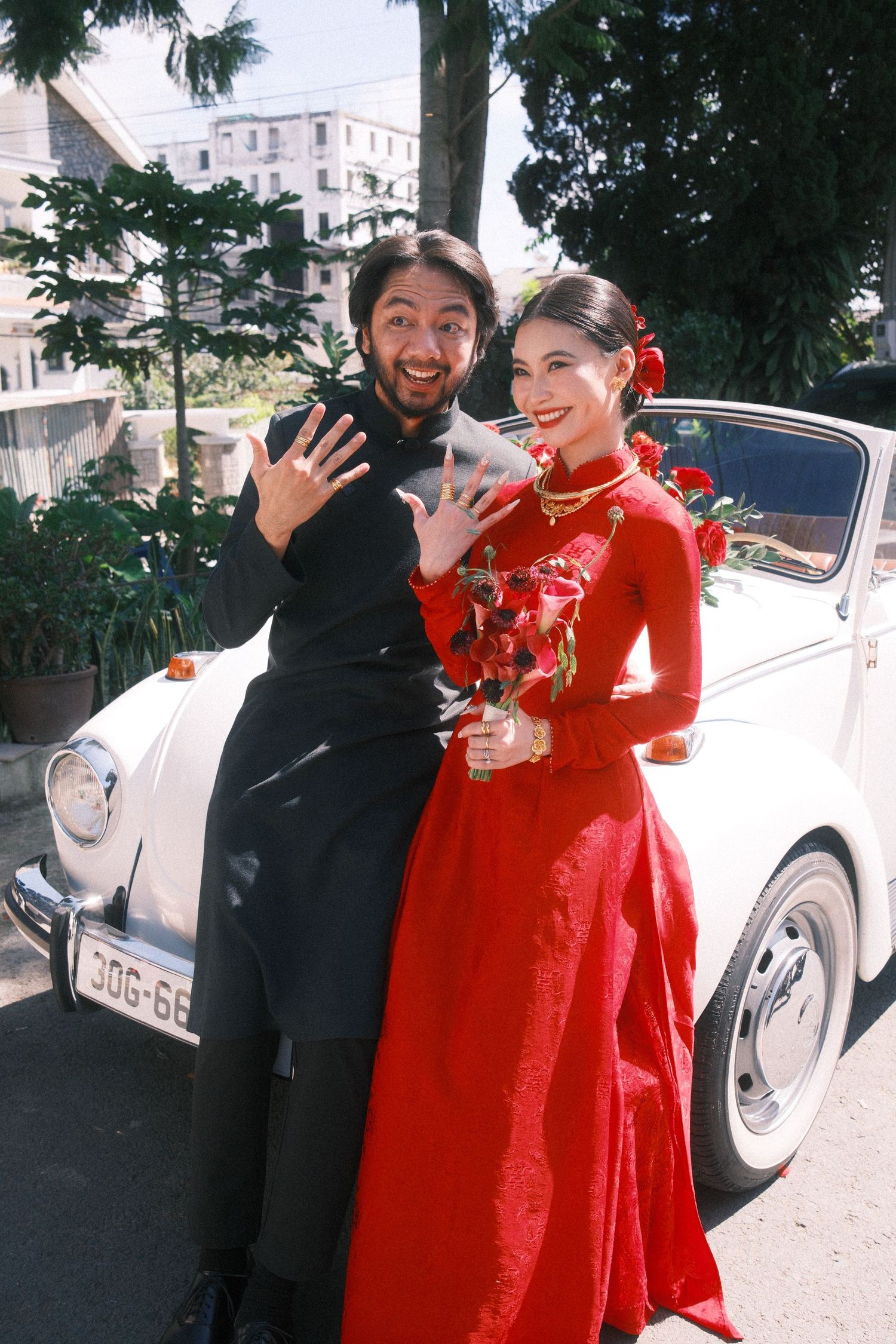 Couple in traditional Vietnamese wedding attire posing by a white vintage car, showing rings.