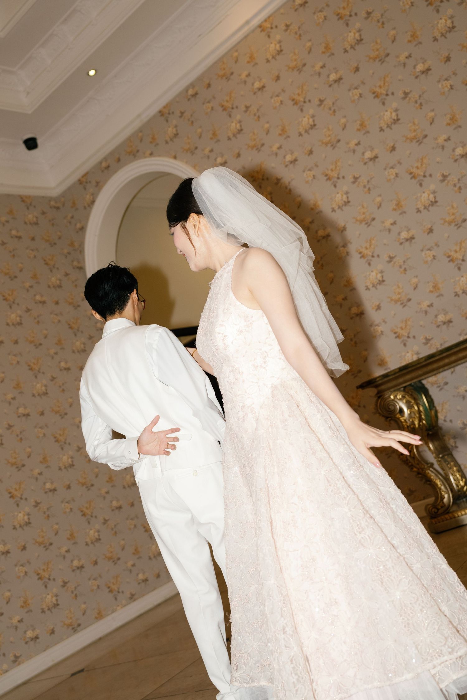 Bride in lace gown and veil dancing with groom in white suit at elegant wedding reception.