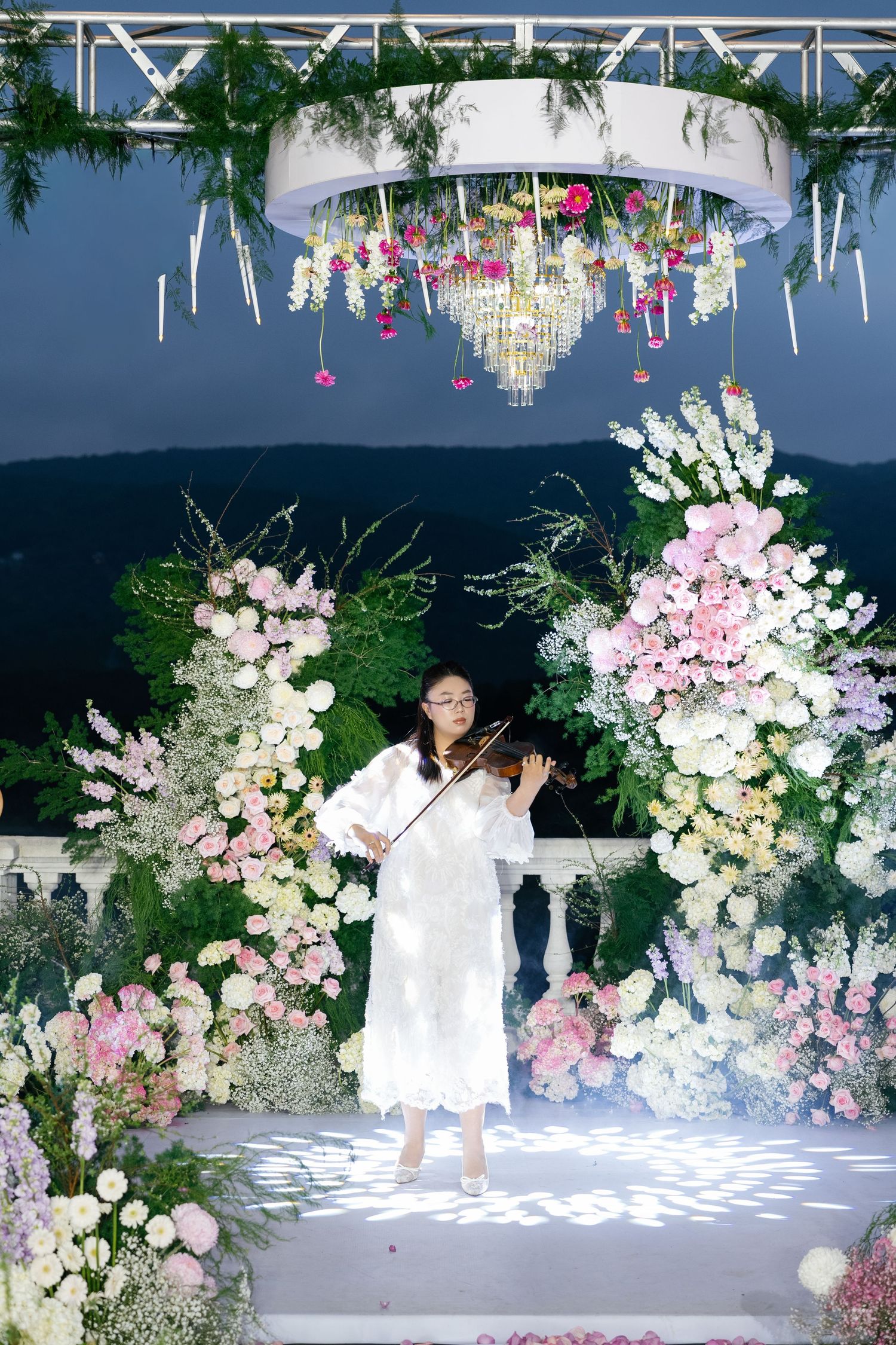 Young violinist in white dress performs at floral wedding arch with pink and white blooms and chandelier.