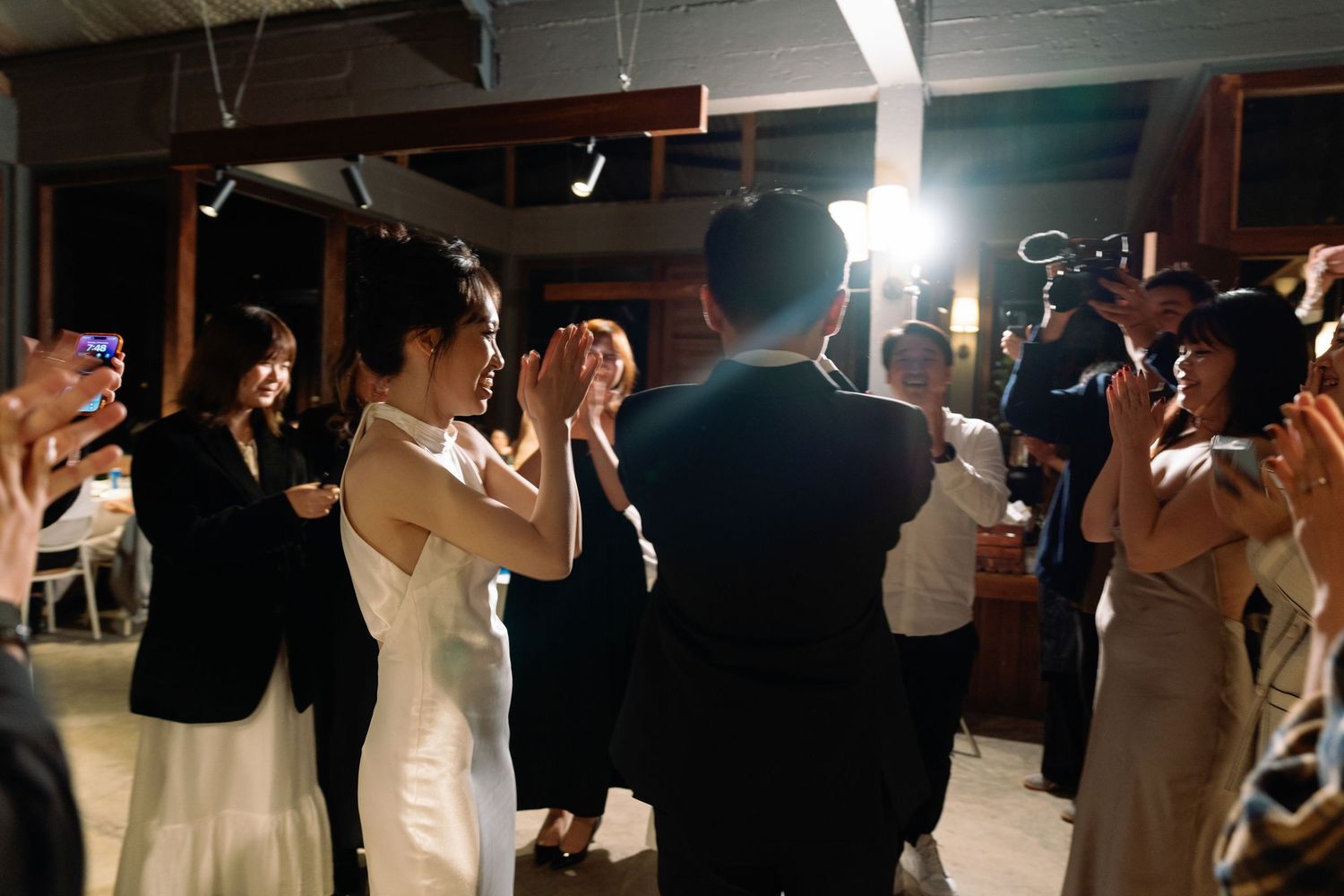 Bride and groom sharing first dance surrounded by applauding guests in a warmly lit wedding reception.