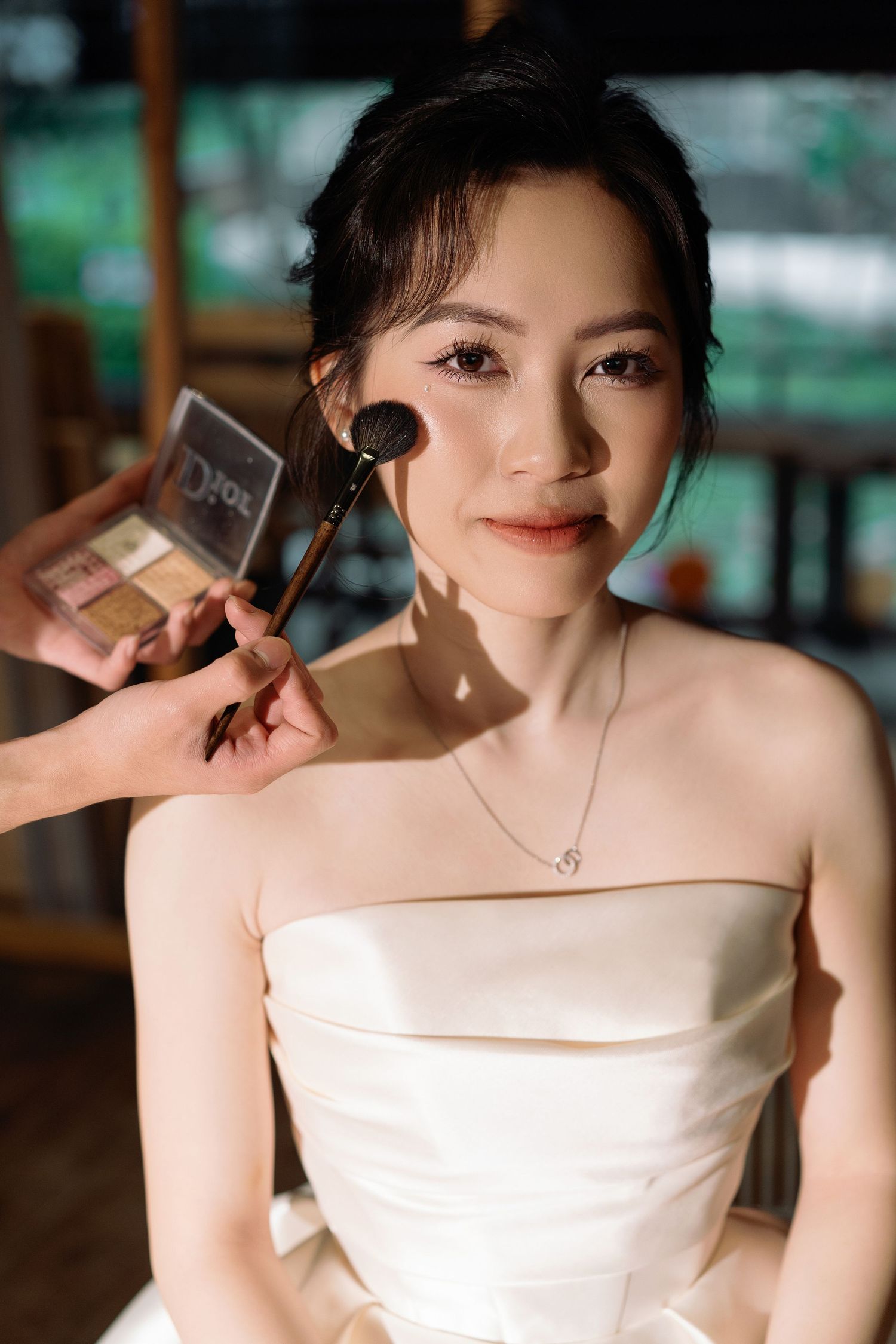 Asian bride in strapless white gown getting makeup applied with a brush during wedding day preparations indoors.