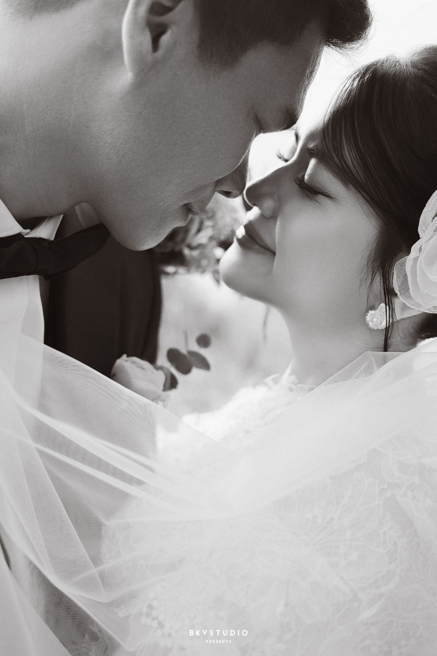 Black and white romantic close-up of bride and groom almost kissing, wrapped in bridal veil on wedding day.