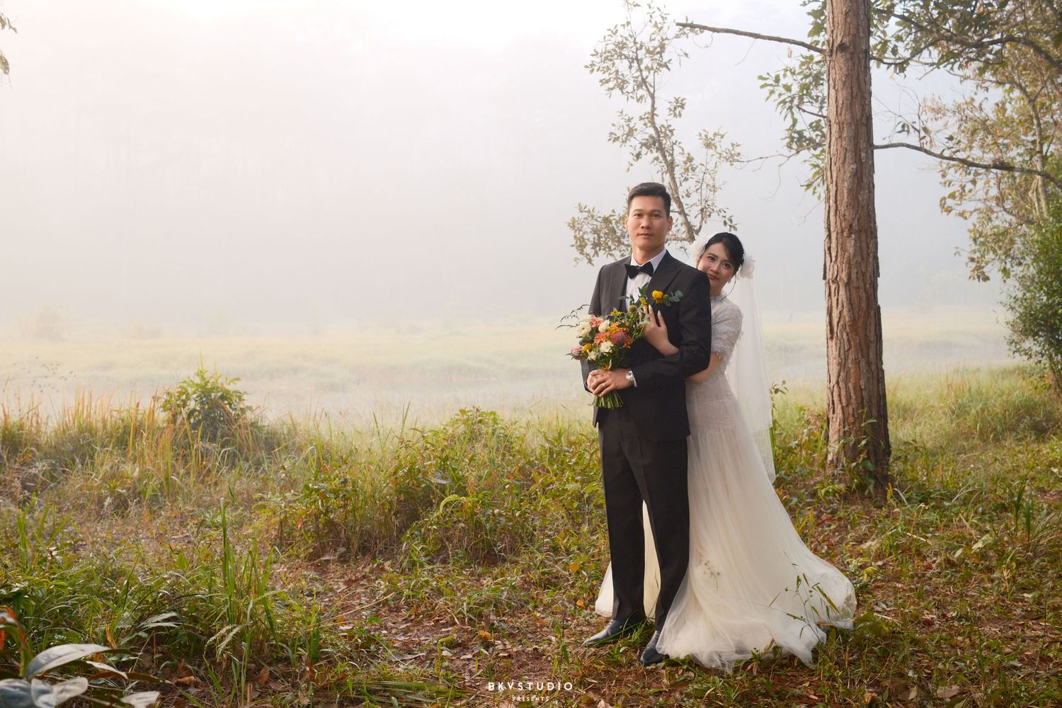 Bride and groom pose together near a pine tree in a misty outdoor landscape during their wedding photoshoot.