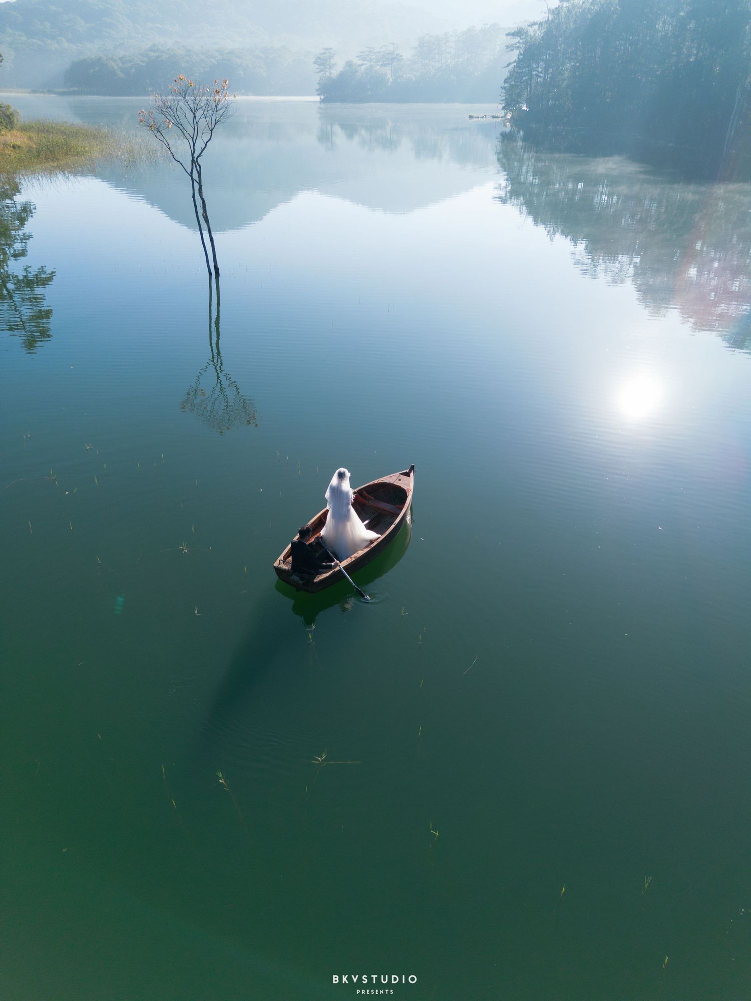 Aerial view of bride in white gown sitting in a small wooden boat on a calm green reflective lake.