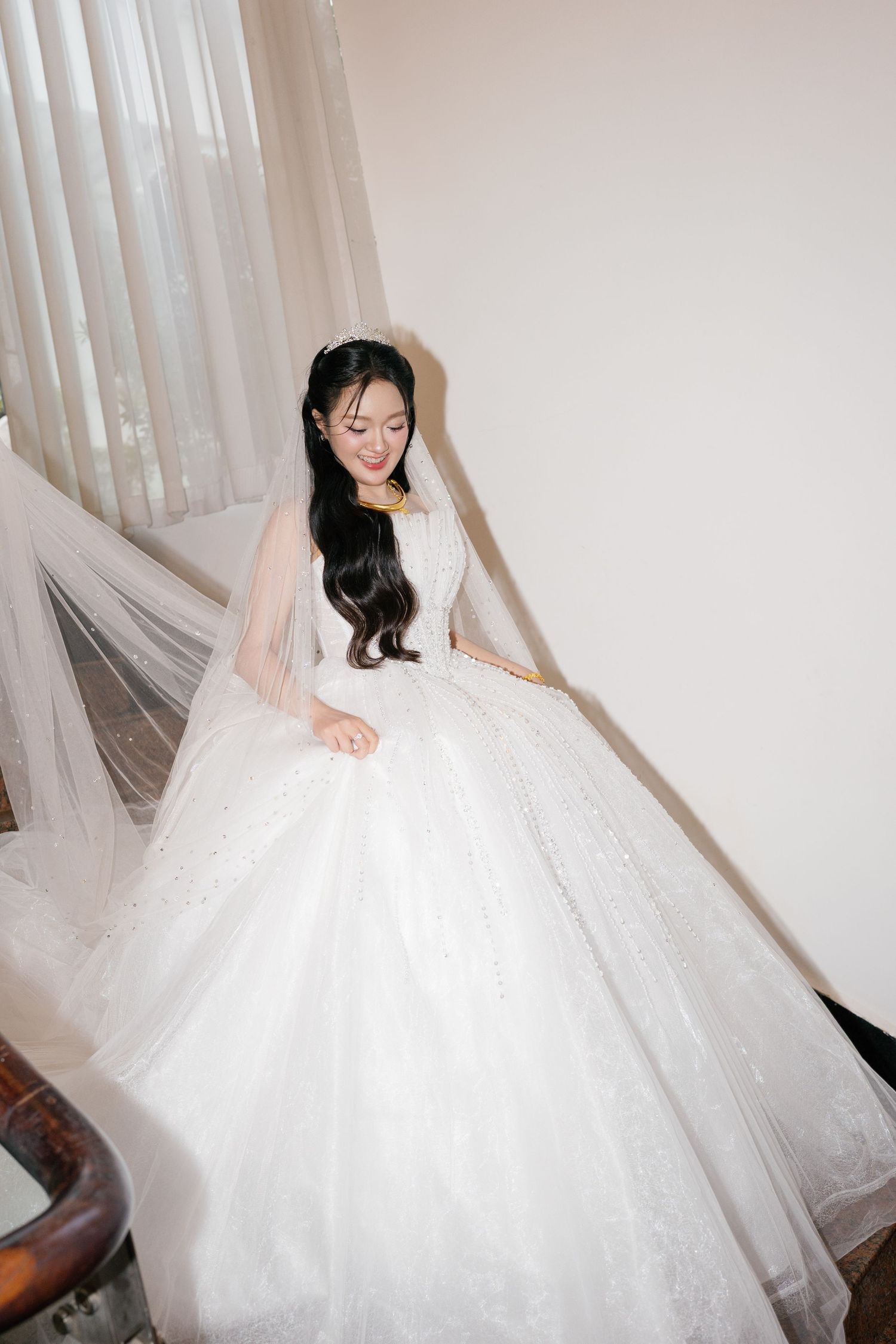 Elegant bride in voluminous white ball gown wedding dress standing on staircase in bright room.