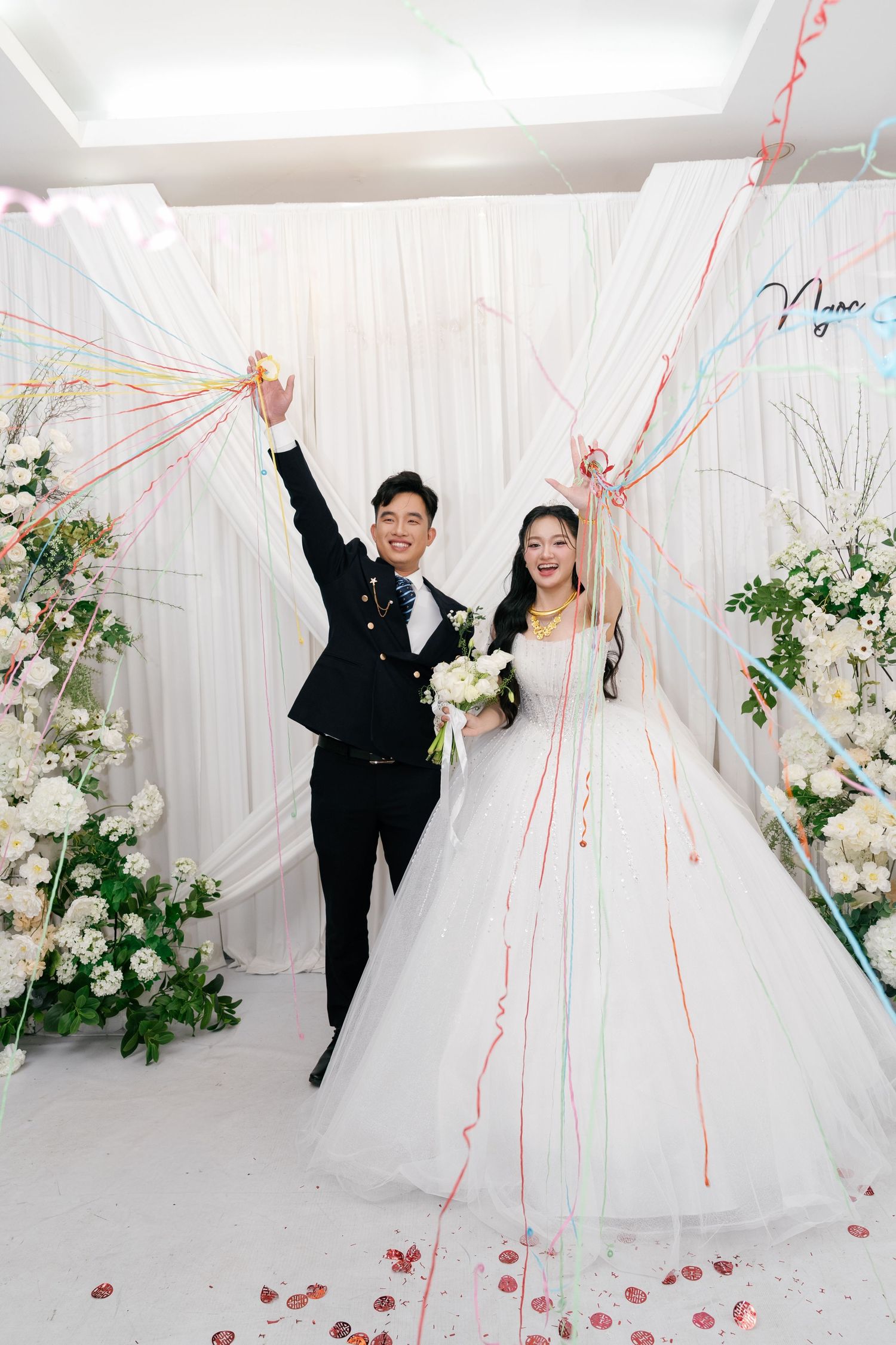 Happy Vietnamese newlyweds celebrate with colorful streamers in white floral decorated wedding venue.