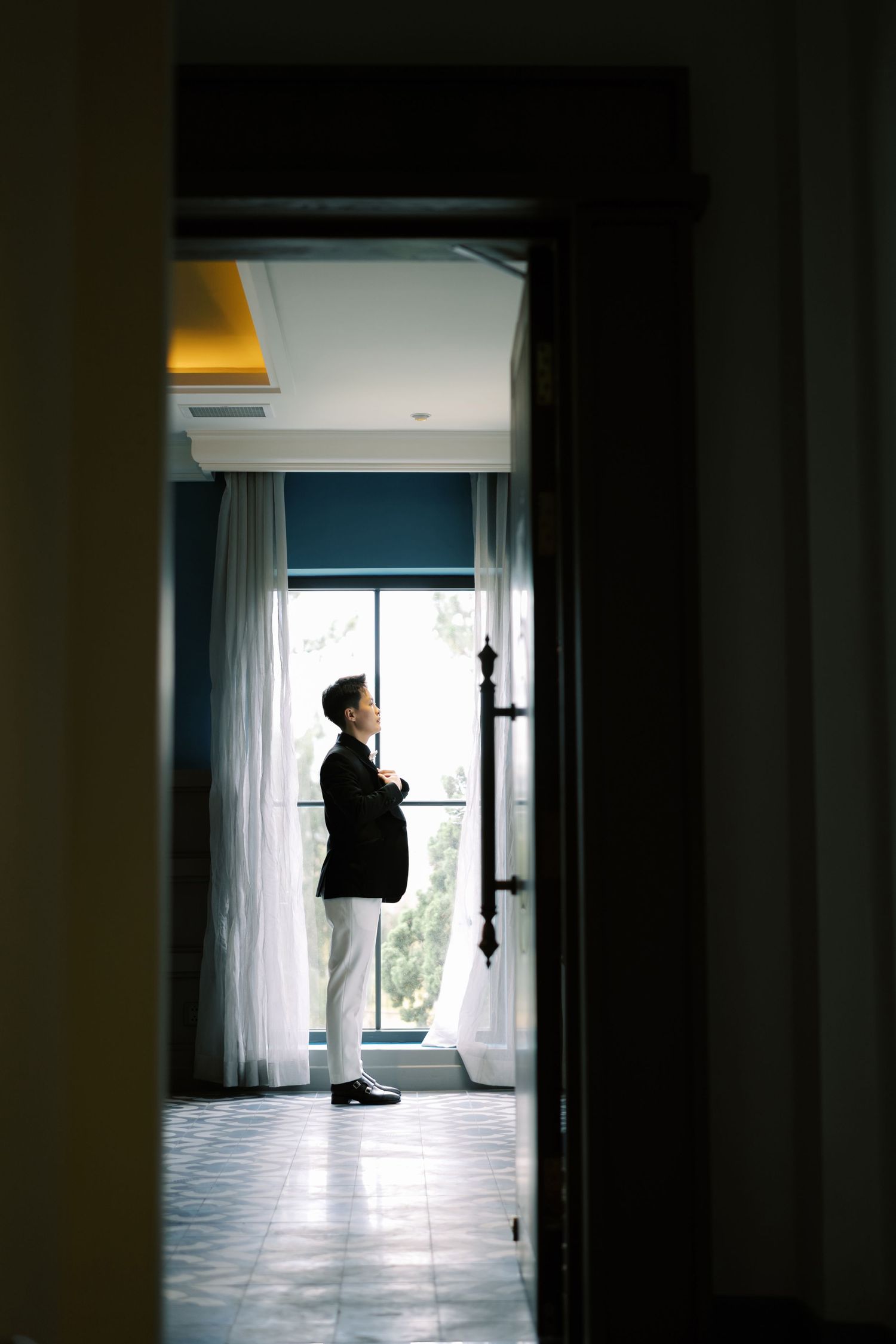 Elegant groom in black suit standing by window in luxury hotel room on wedding day.