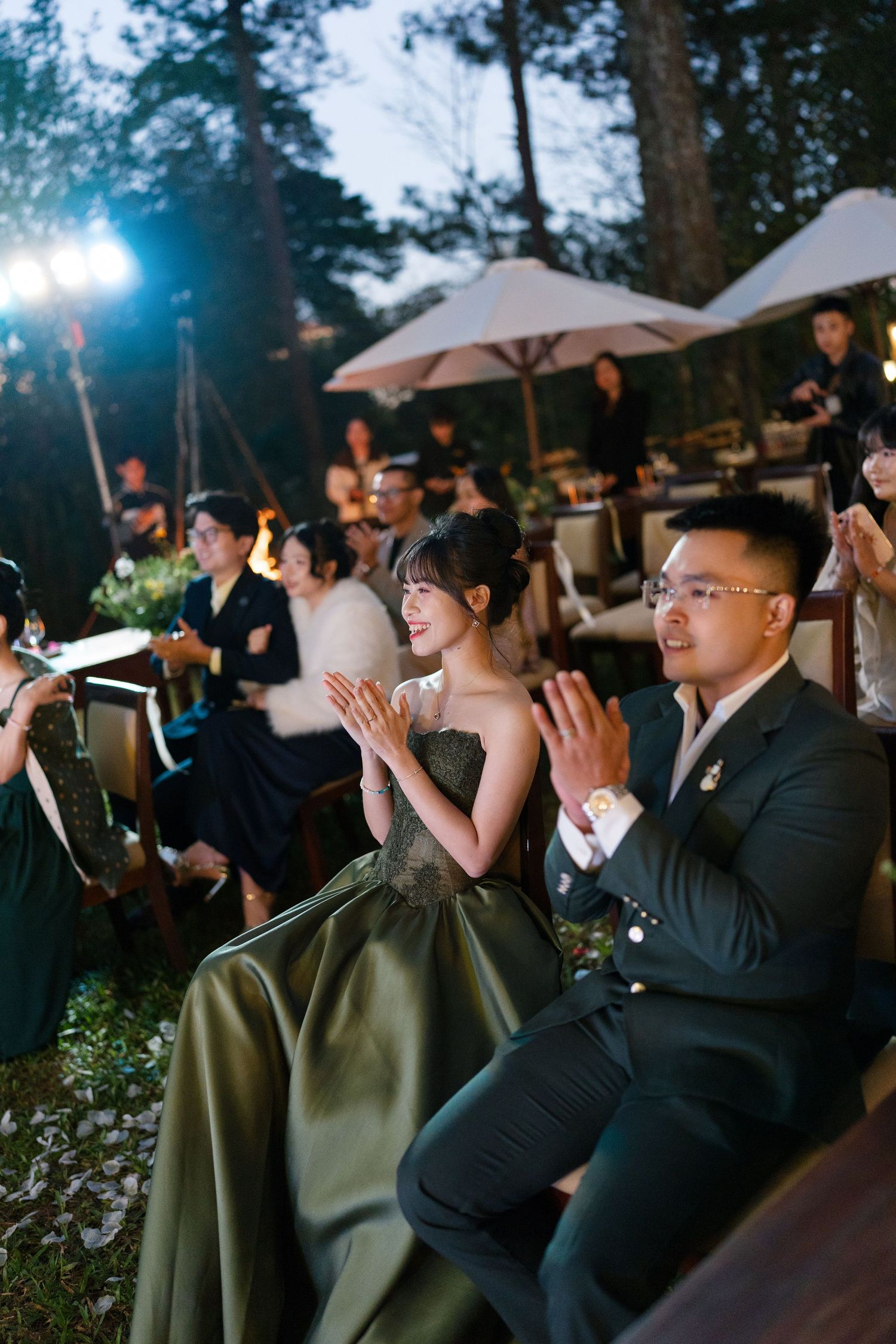 Wedding guests in formal attire clapping and smiling at outdoor evening reception in forest setting.