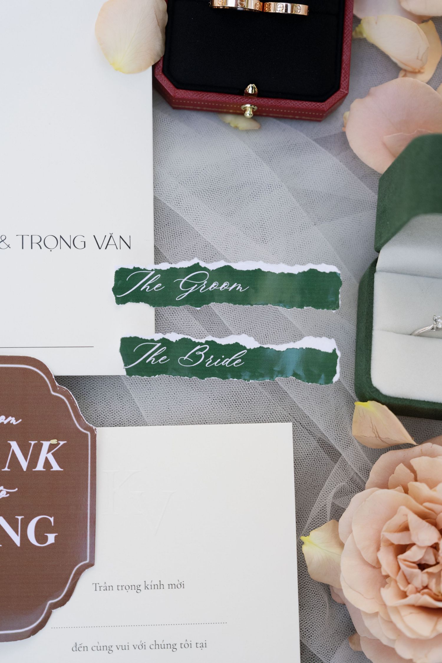 Wedding stationery flat lay with green bride and groom labels, rings, and pink roses.