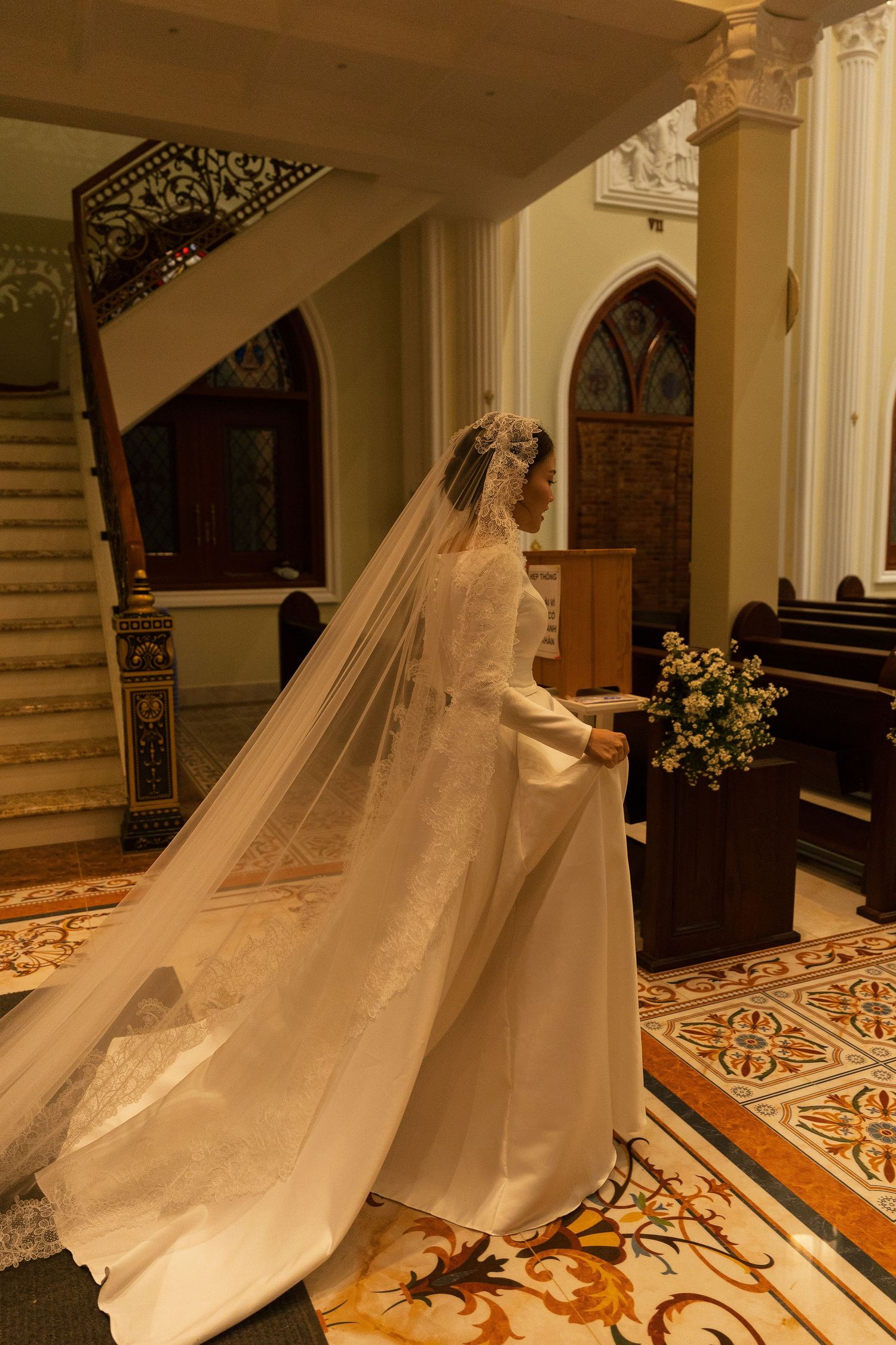 Bride in elegant long-sleeve wedding gown with flowing cathedral veil standing inside ornate church with marble floors.