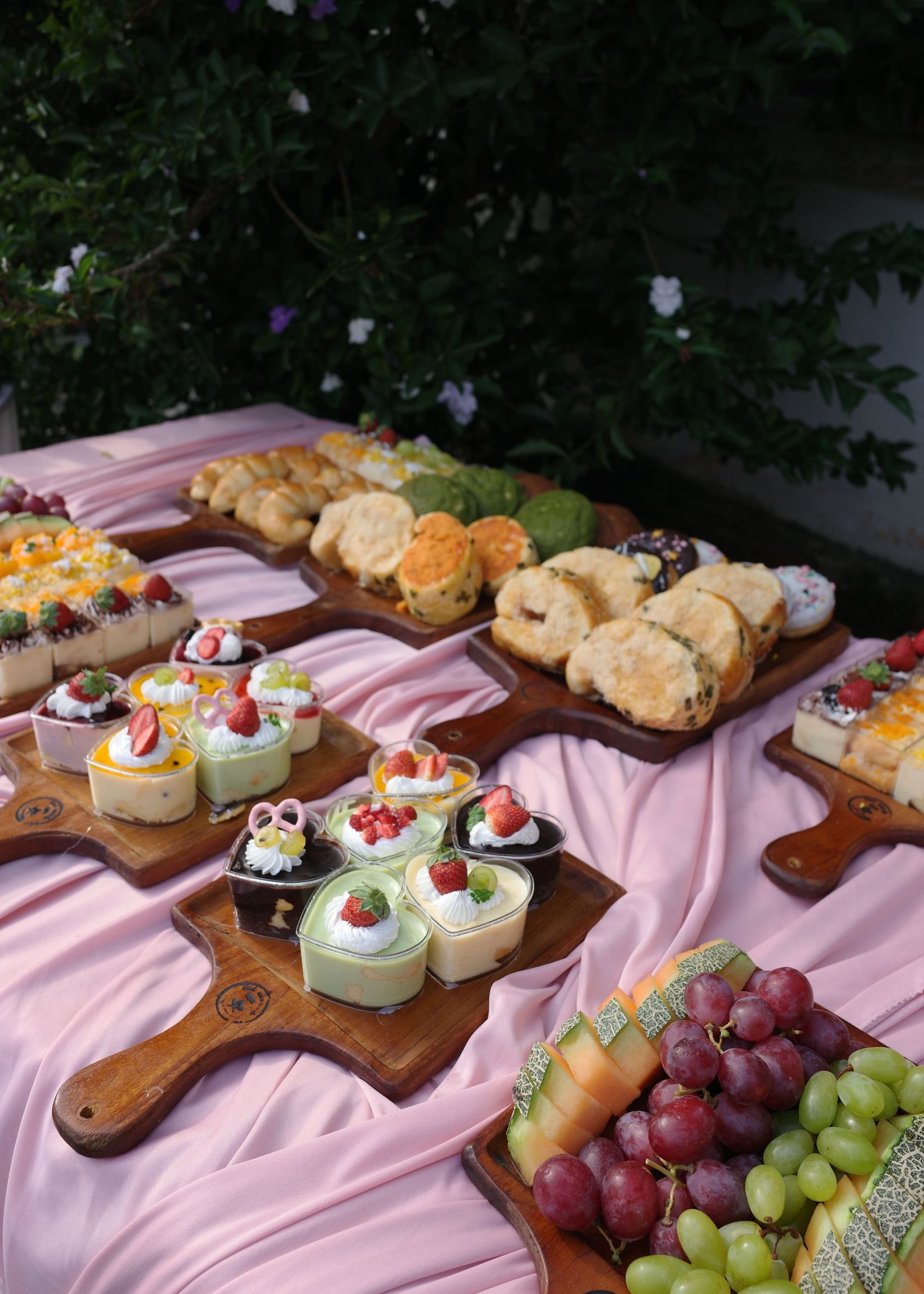 Elegant dessert table with assorted mini cakes, pastries, and fresh grapes on wooden boards with pink tablecloth.