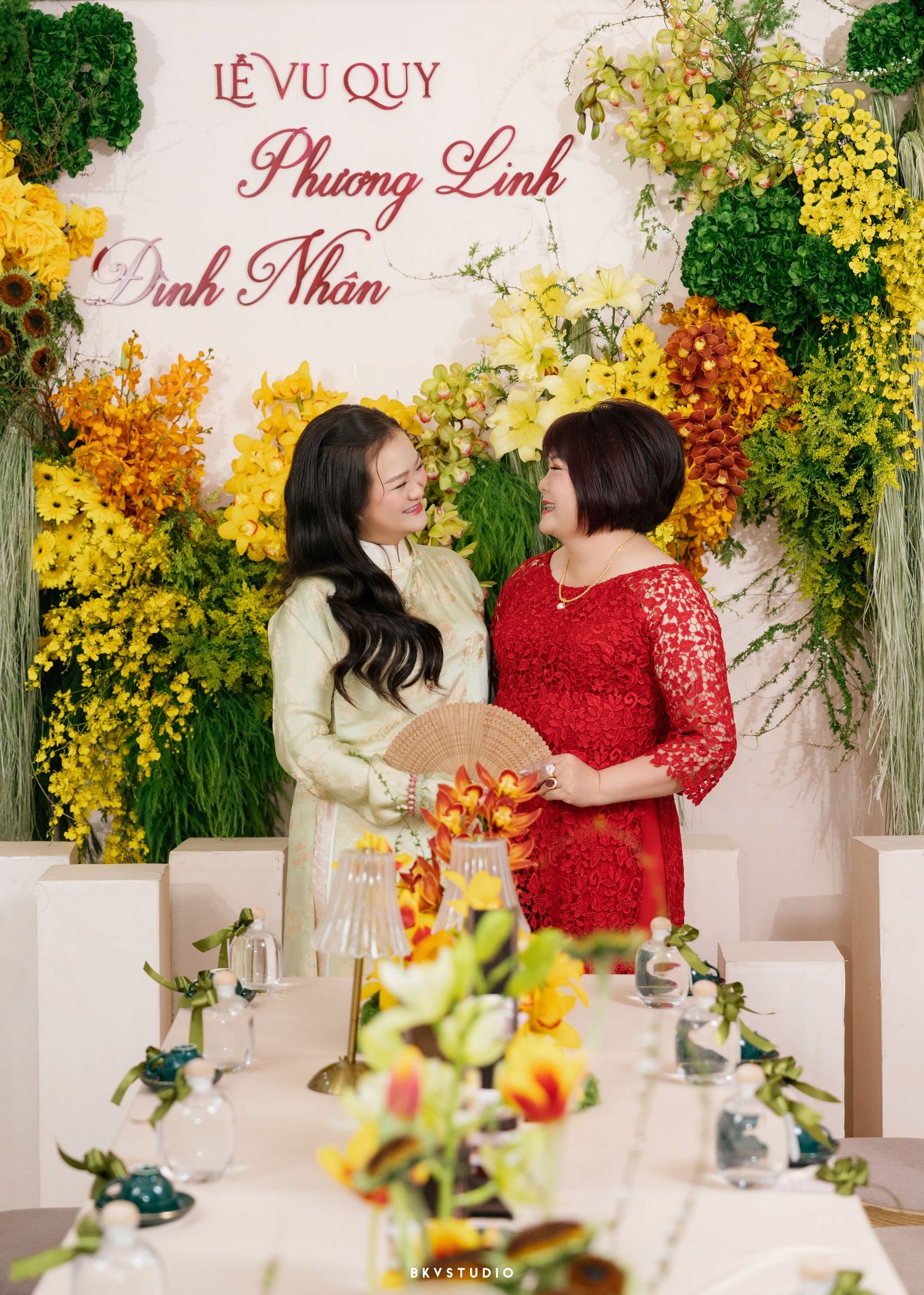 Two women in traditional Vietnamese attire smile at a Le Vu Quy wedding ceremony with colorful floral backdrop.