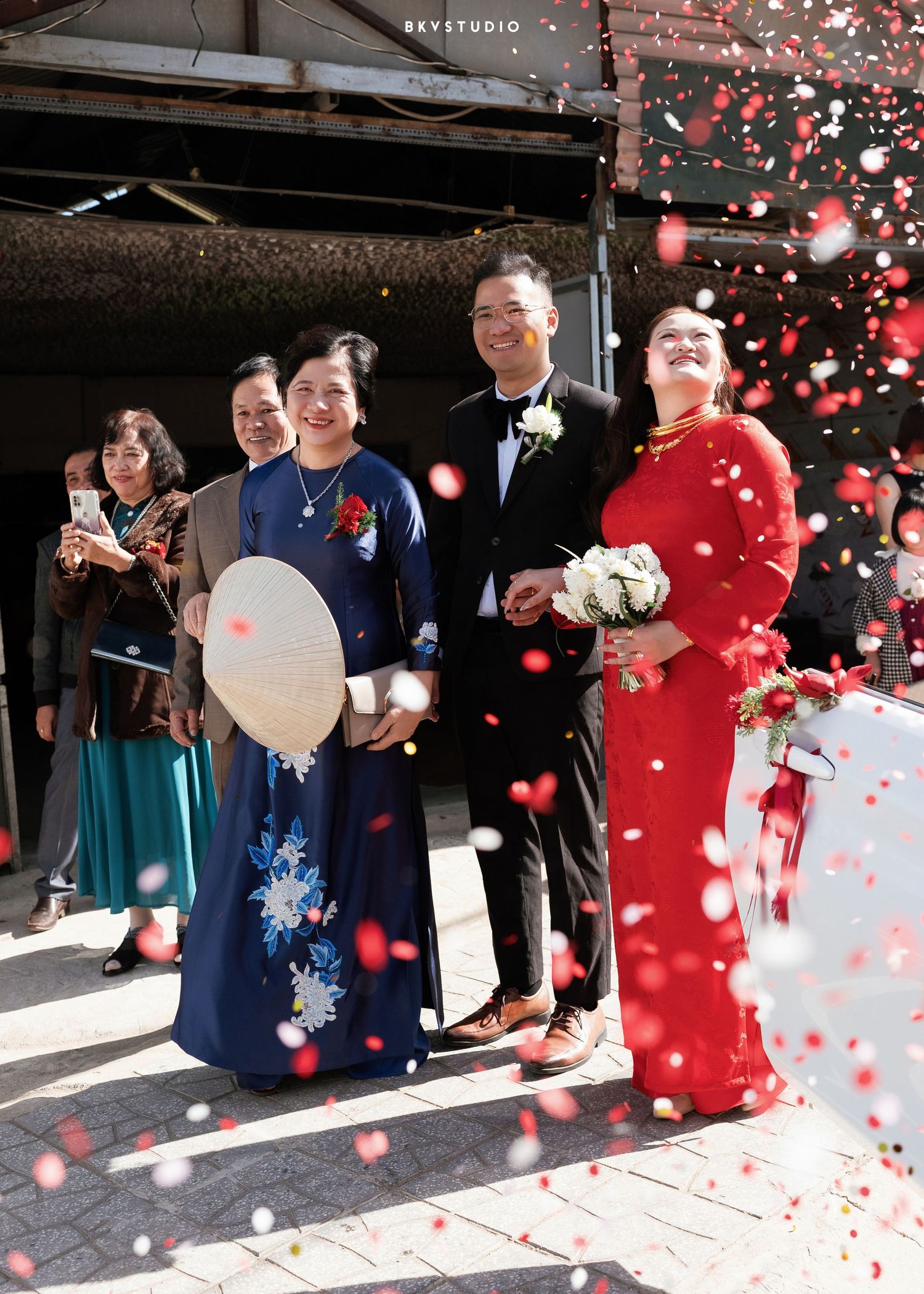 Vietnamese wedding ceremony with bride in red ao dai, groom in tuxedo, and guests celebrating with confetti.