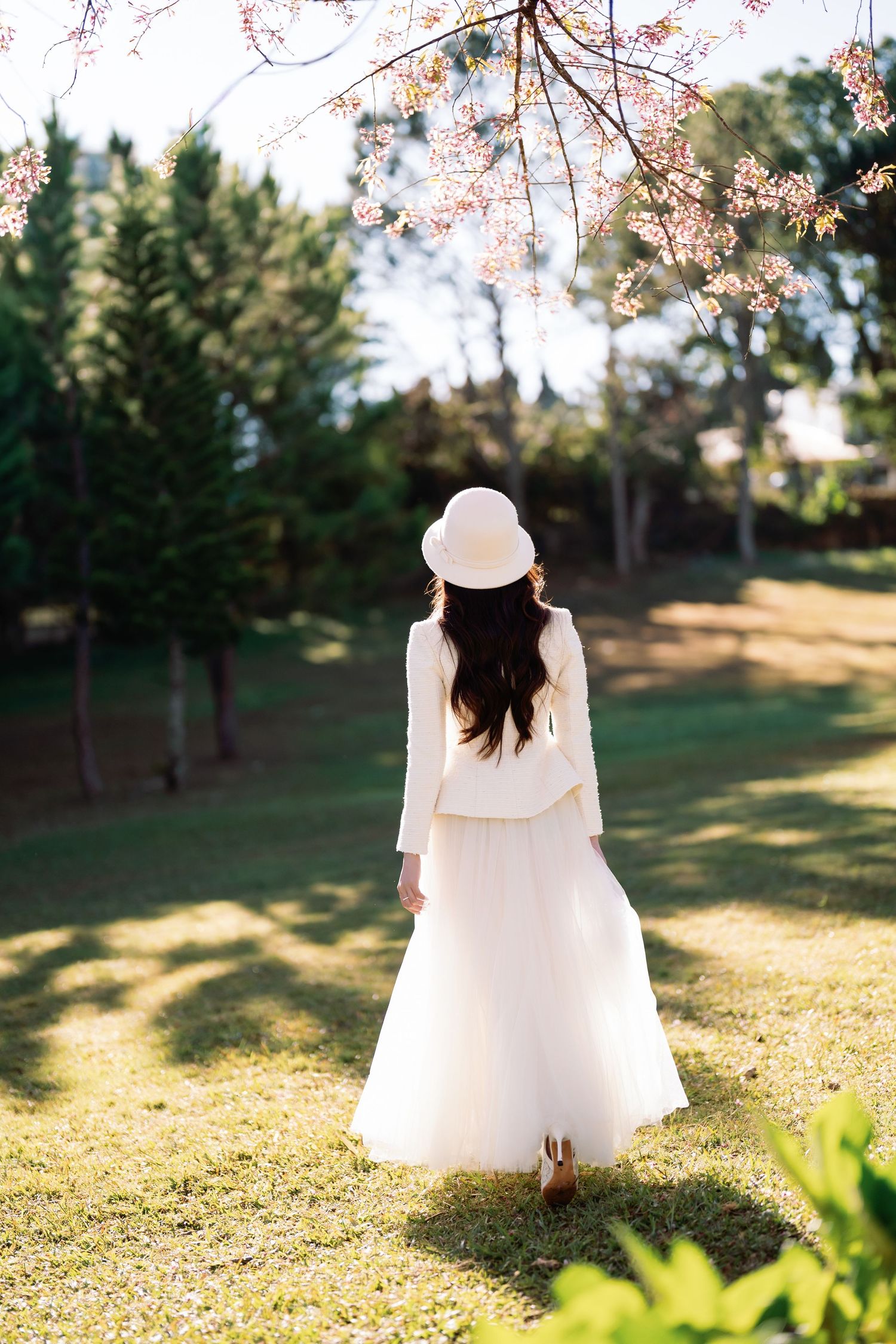 Woman in white dress and hat stands in sunny park under blooming cherry blossom tree.