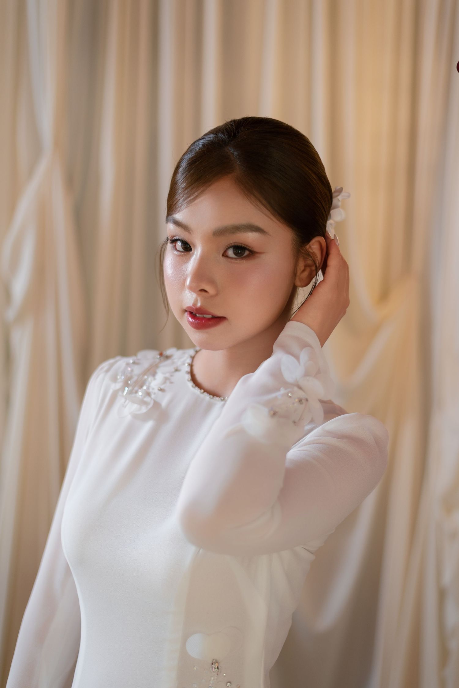 Young Asian woman in white dress with floral embroidery, posing gracefully with hand near hair against draped curtains.