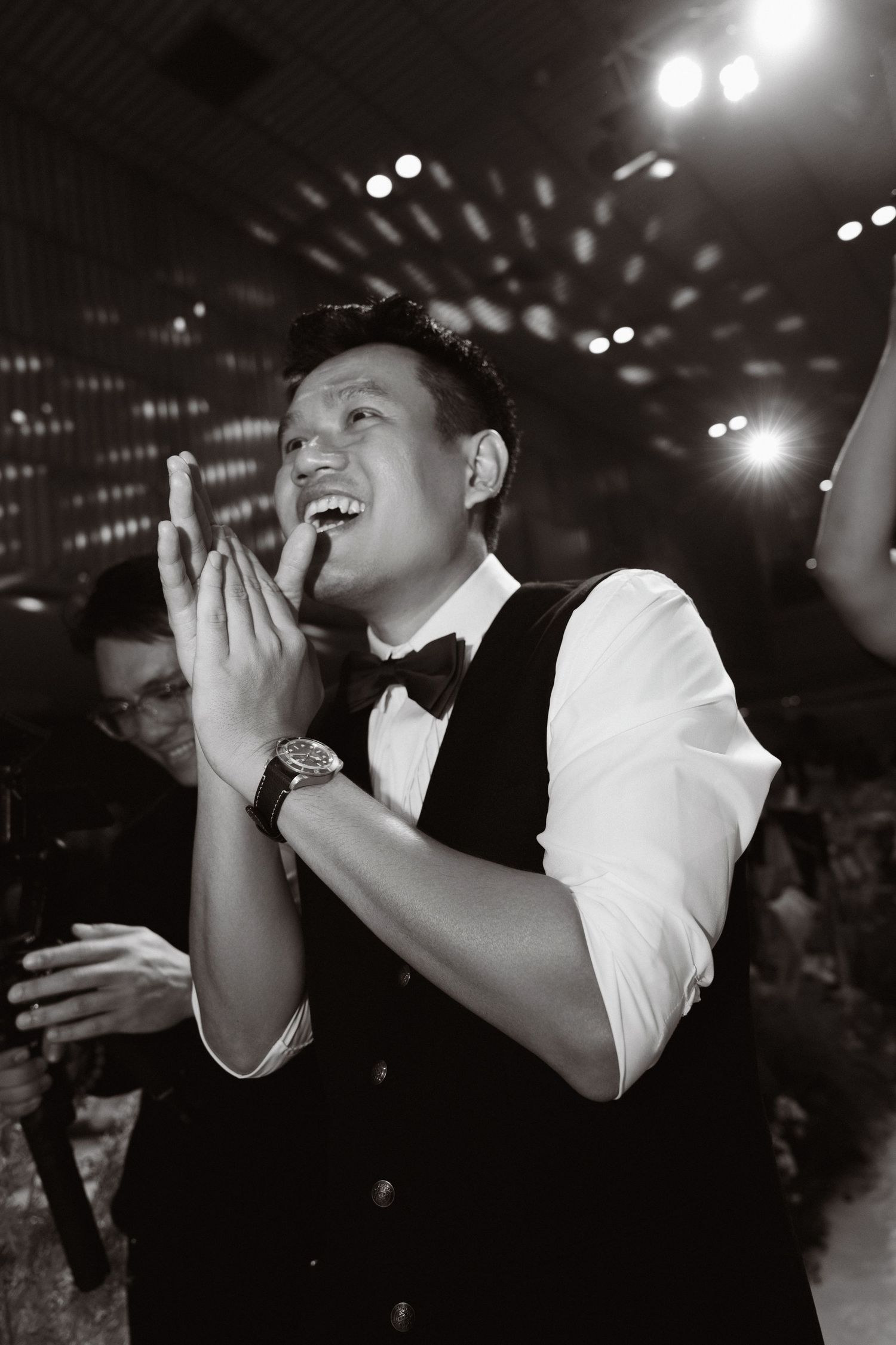 Black and white photo of a smiling man in a vest and bow tie clapping at a formal event.