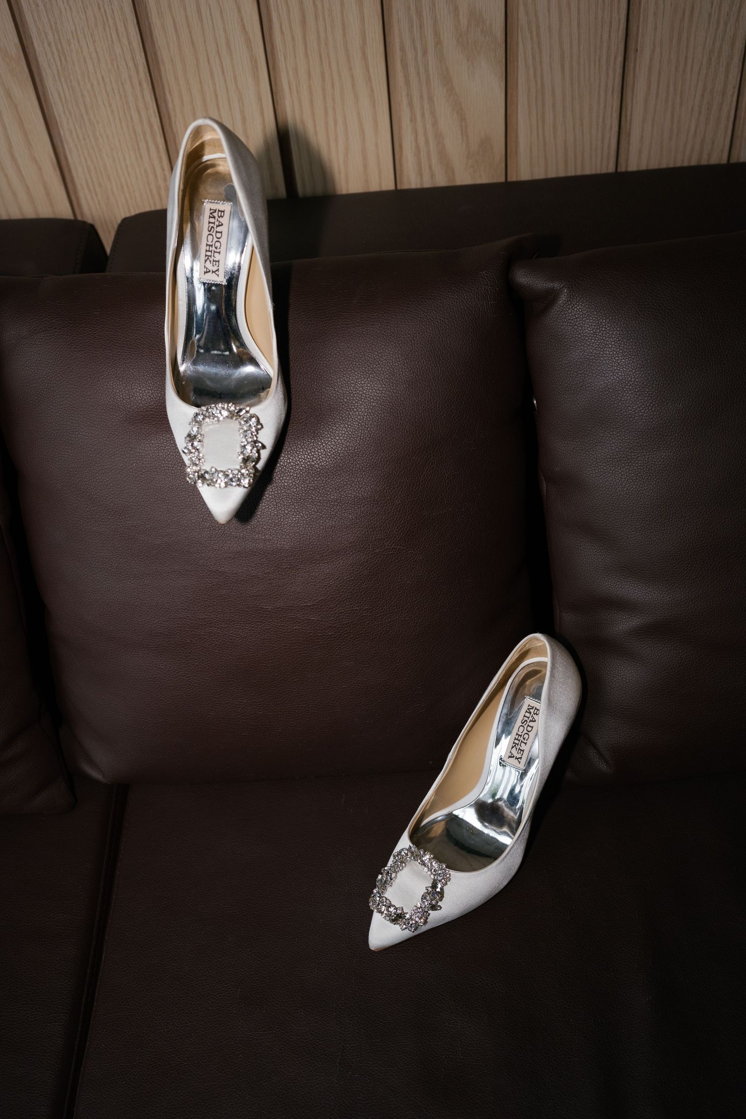 White bridal heels with crystal buckle embellishment displayed on dark brown leather sofa cushions.