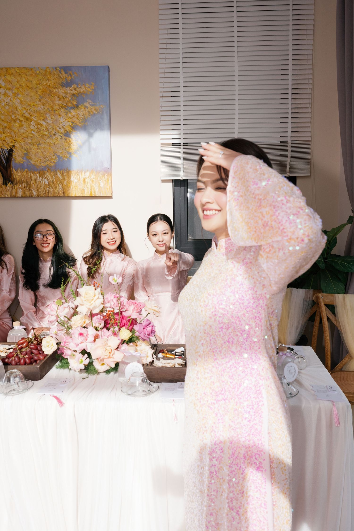 Smiling bride in sparkly pink ao dai poses while bridesmaids gather around flower-decorated table.