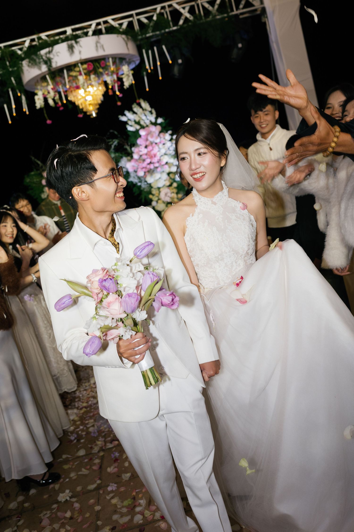 Asian couple in white wedding attire walking down the aisle at night, bride holding purple tulip bouquet.