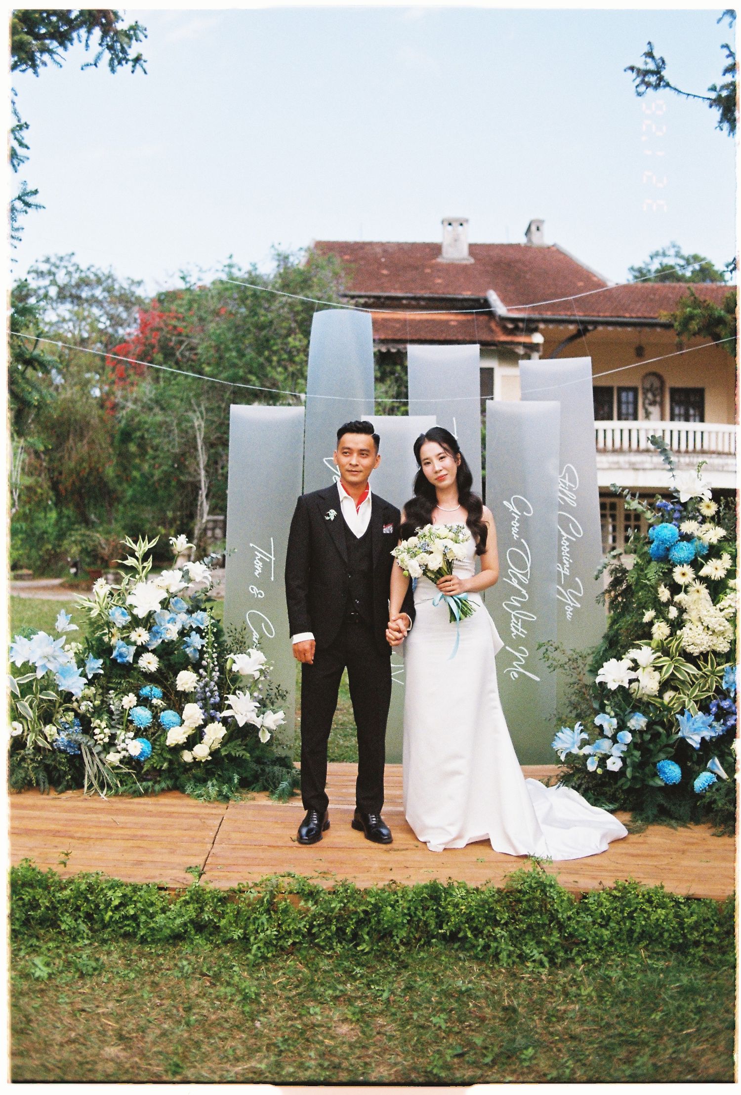 Couple poses in wedding attire before floral backdrop with blue and white flowers at outdoor garden ceremony.