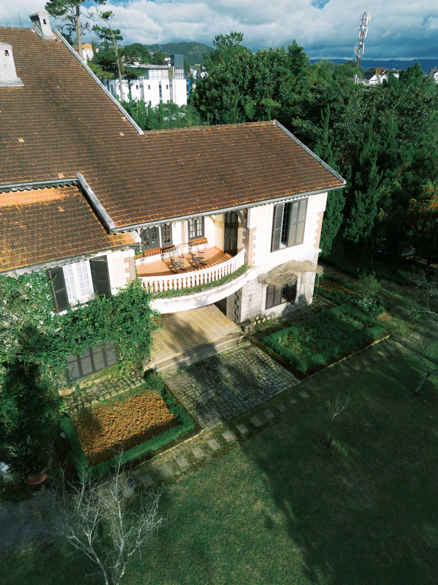 Aerial view of a charming two-story villa with terracotta roof, white walls, balcony, and manicured gardens surrounded by lush trees.