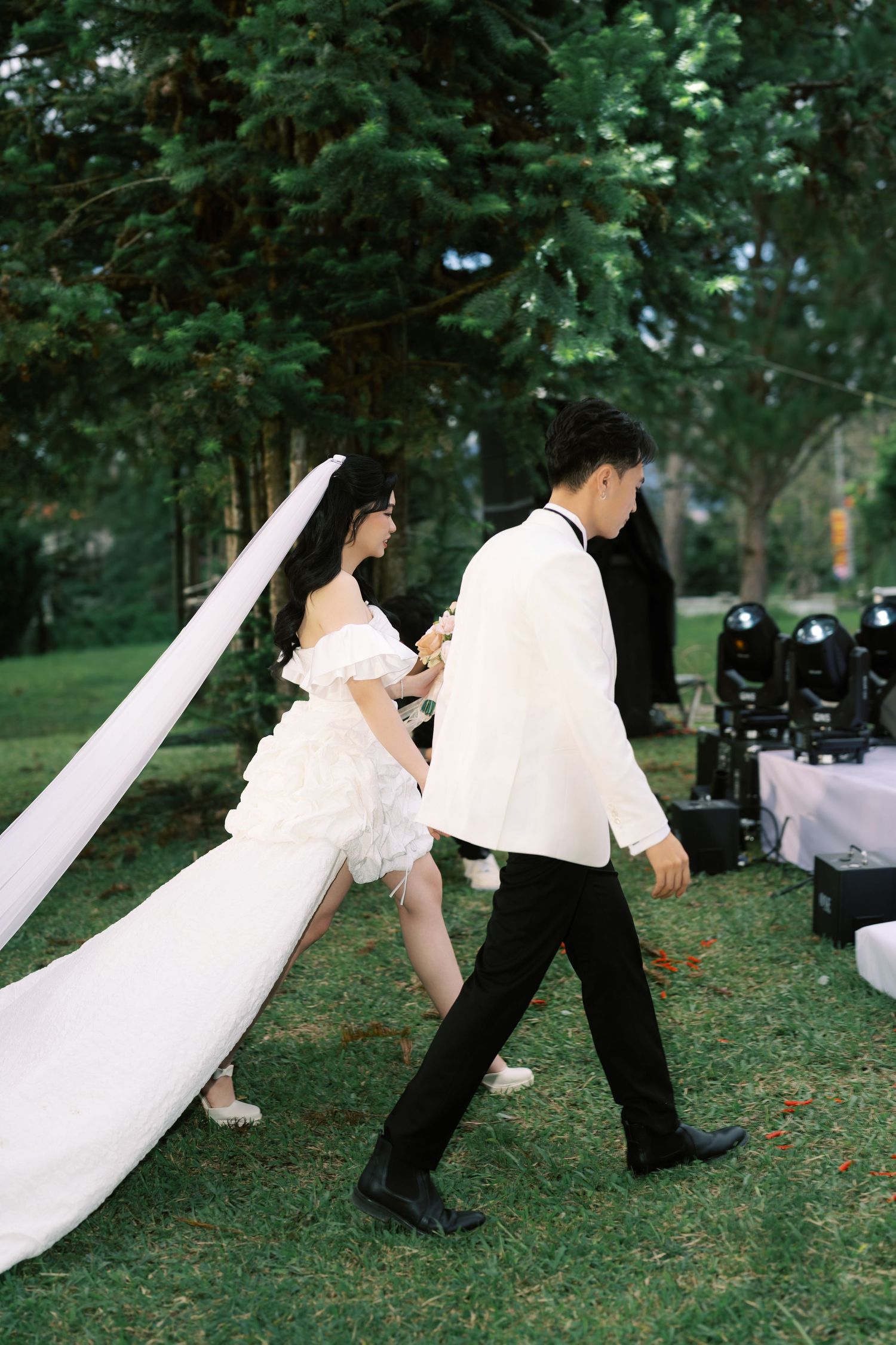 Bride in white gown and groom in white suit walking outdoors on green grass during wedding ceremony.