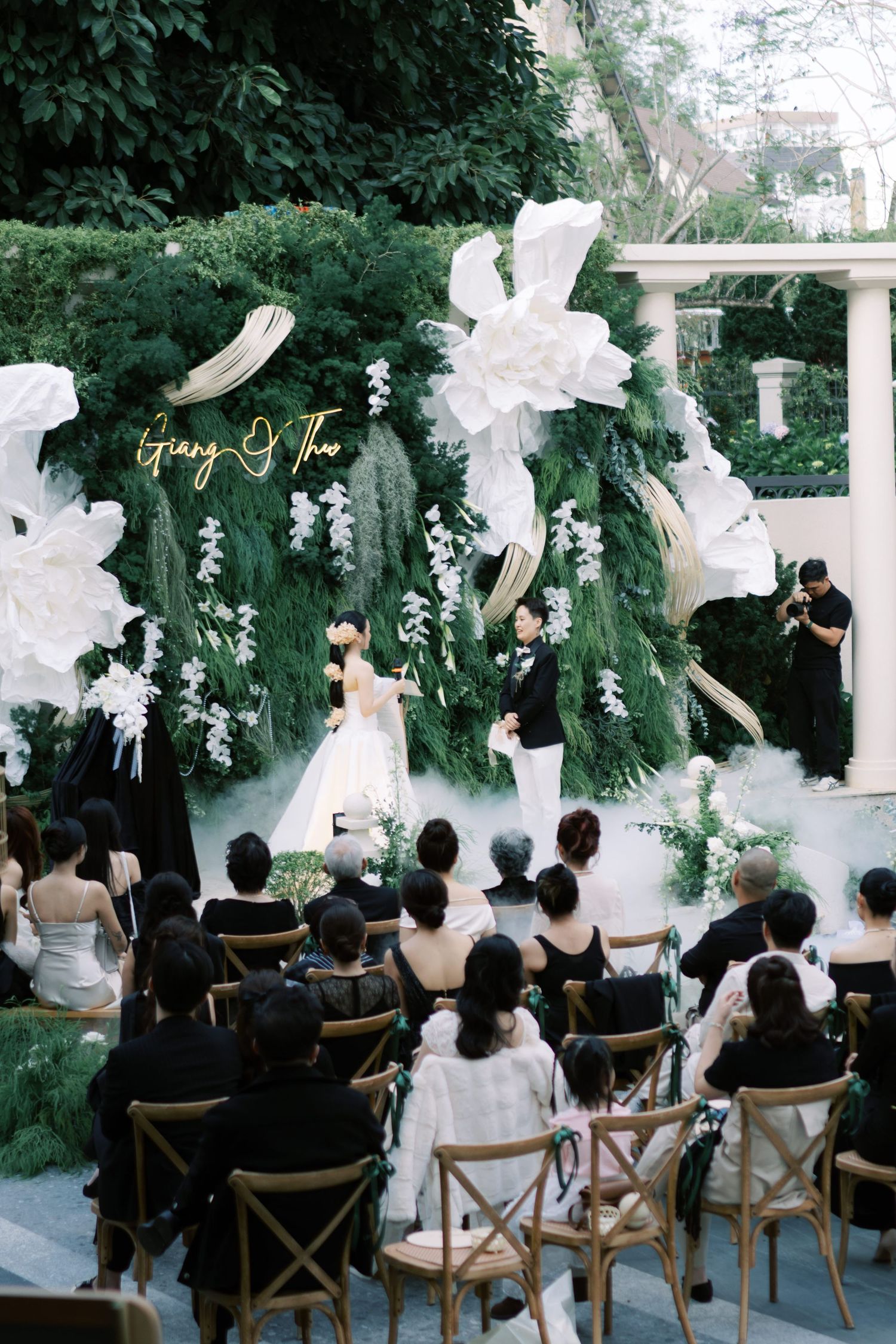 Outdoor wedding ceremony with guests seated before lush green floral backdrop and misty atmosphere.