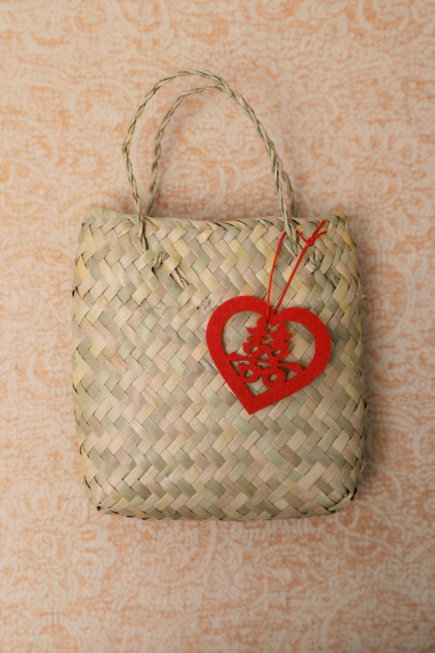 Woven straw tote bag with red heart-shaped double happiness charm on floral background.