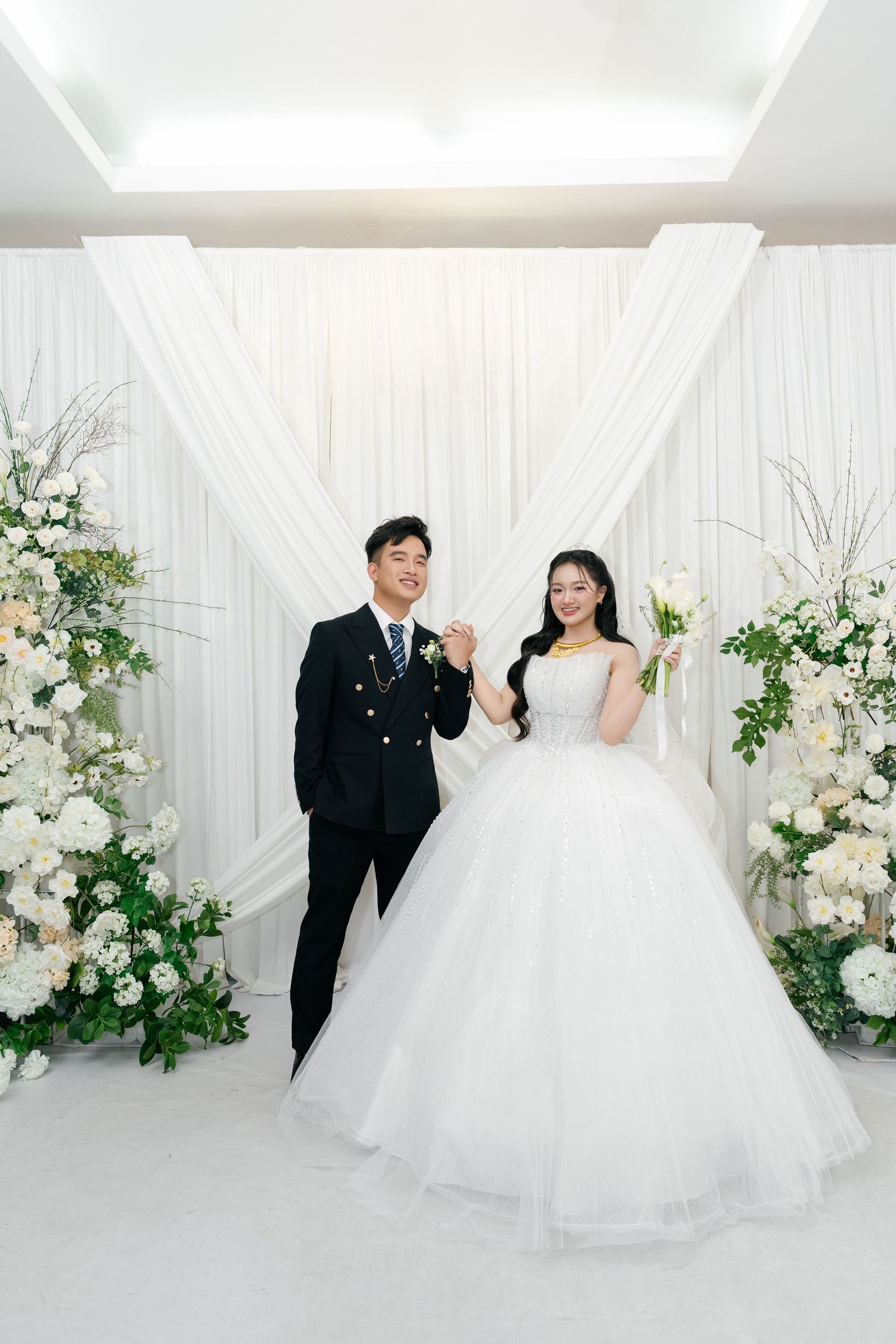 A joyful couple poses in wedding attire before a white draped backdrop adorned with elegant floral arrangements.