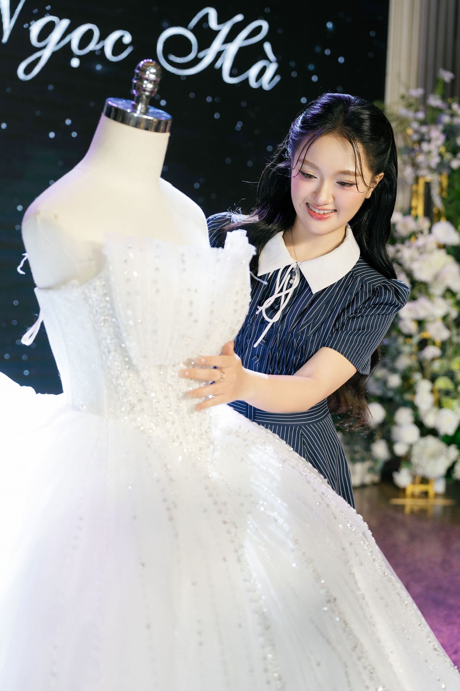 Smiling young woman in striped dress admires ornate white ballgown wedding dress on mannequin.