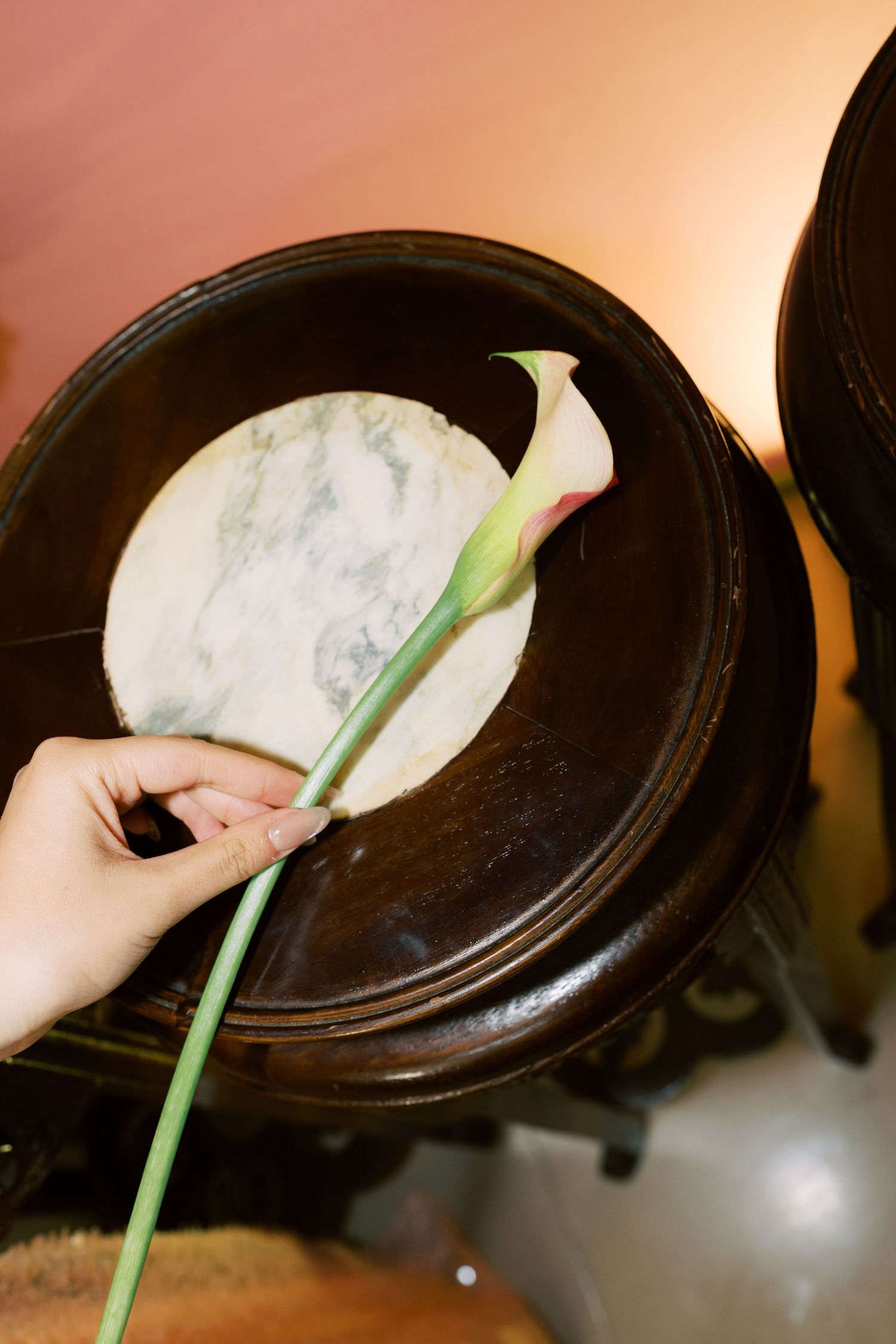 A hand placing a single calla lily flower on a dark wooden decorative bowl with marble insert.