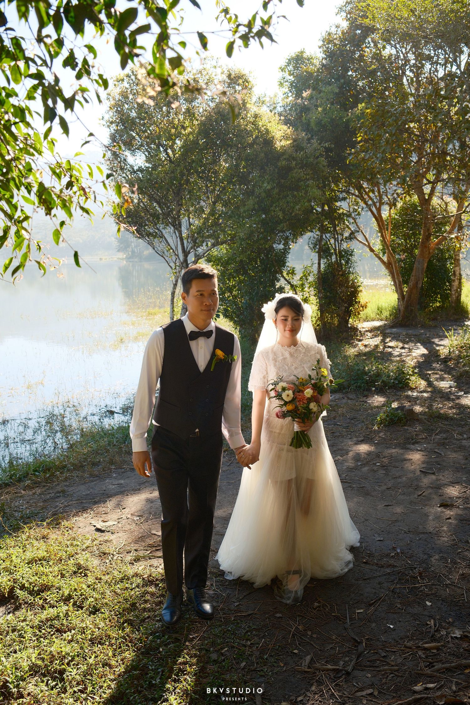 Bride and groom walk hand in hand along a lakeside path in lush green outdoor setting.