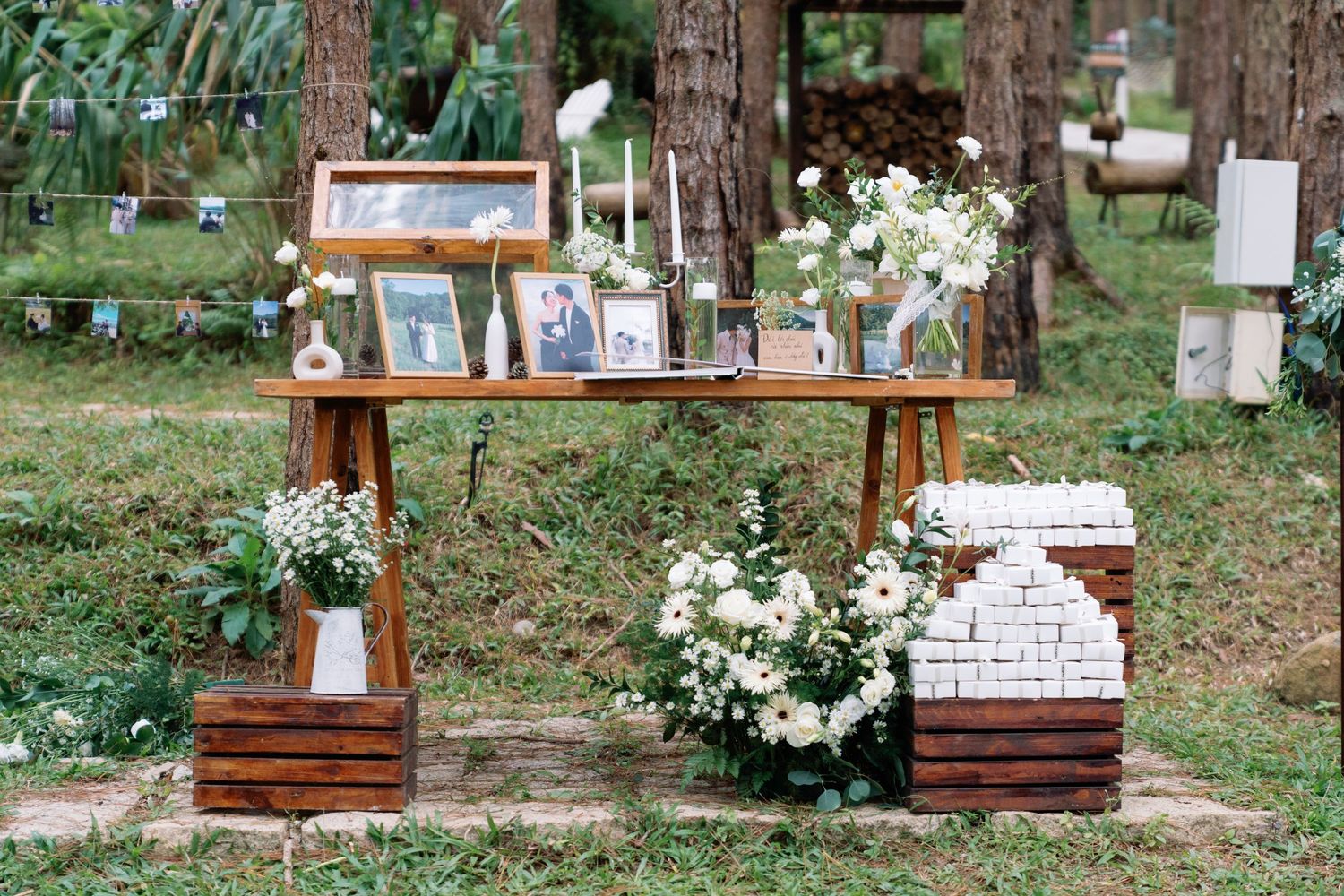 Rustic outdoor wedding display table with white floral arrangements, framed photos, and candles set among pine trees.