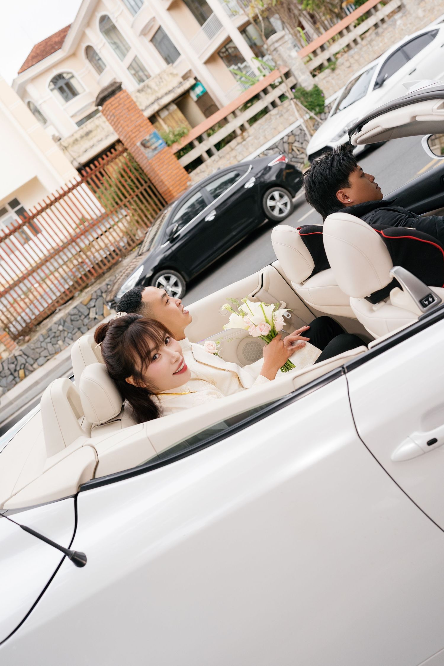 Bride holding bouquet sits in white convertible car with groom during wedding day celebration.