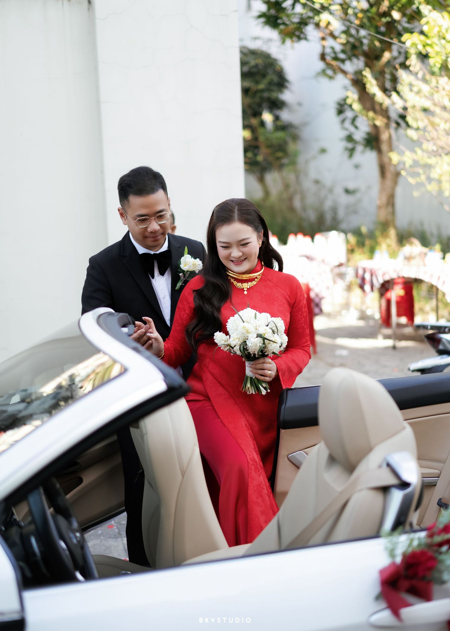 Vietnamese bride in red ao dai holds white bouquet beside groom in tuxedo near white convertible car.