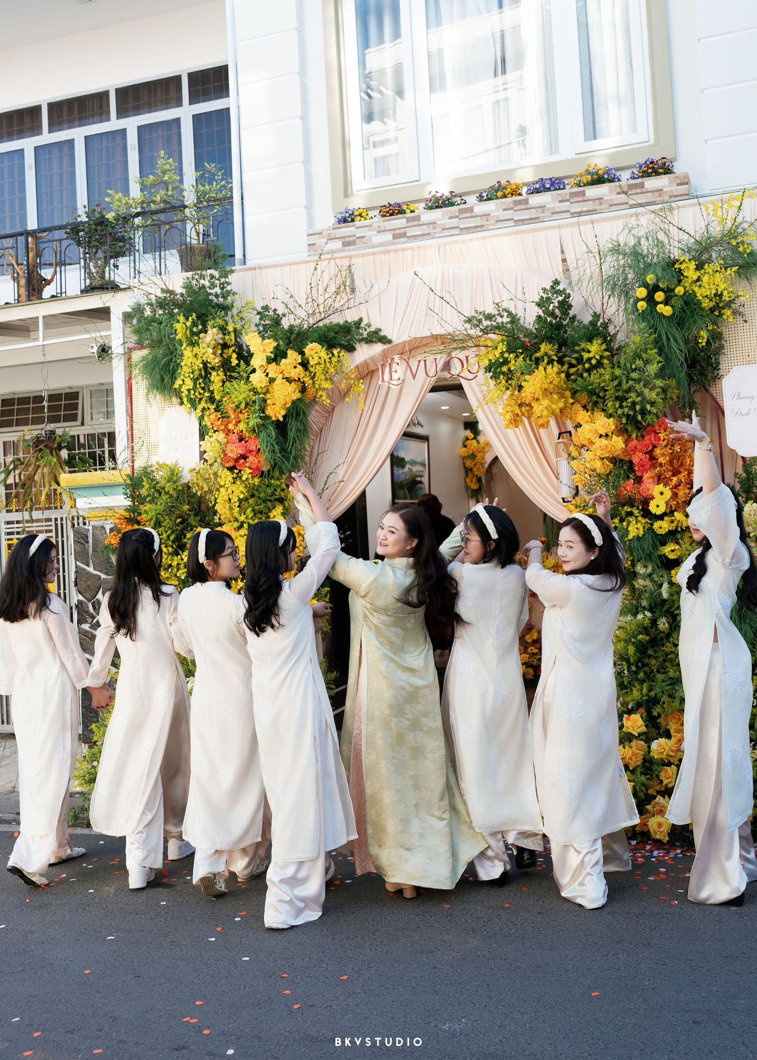 Women in white ao dai dresses gather under a floral arch decorated with yellow and orange blooms at a Vietnamese wedding.