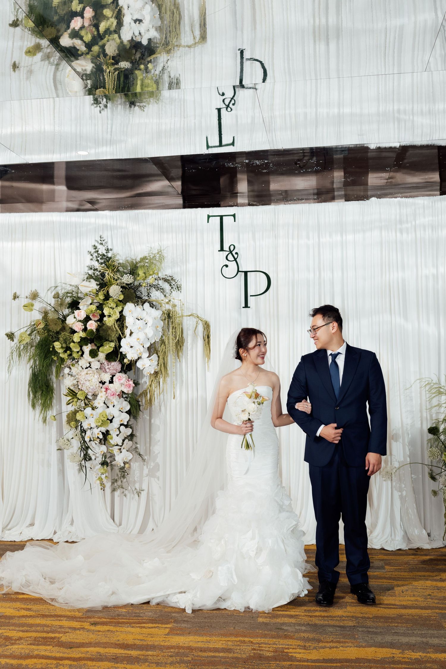 Bride and groom standing together at wedding altar with floral arrangement and monogram backdrop.
