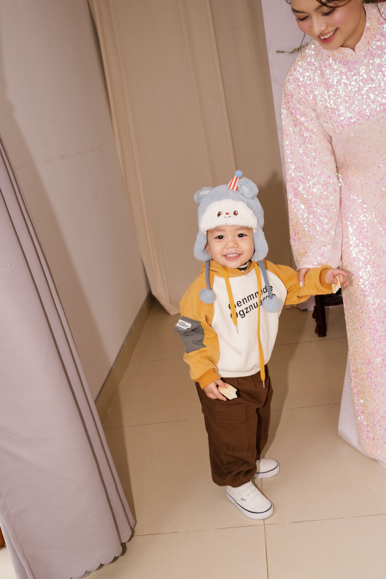 Adorable toddler in bear hat and yellow hoodie smiles while holding hand at Asian wedding celebration.