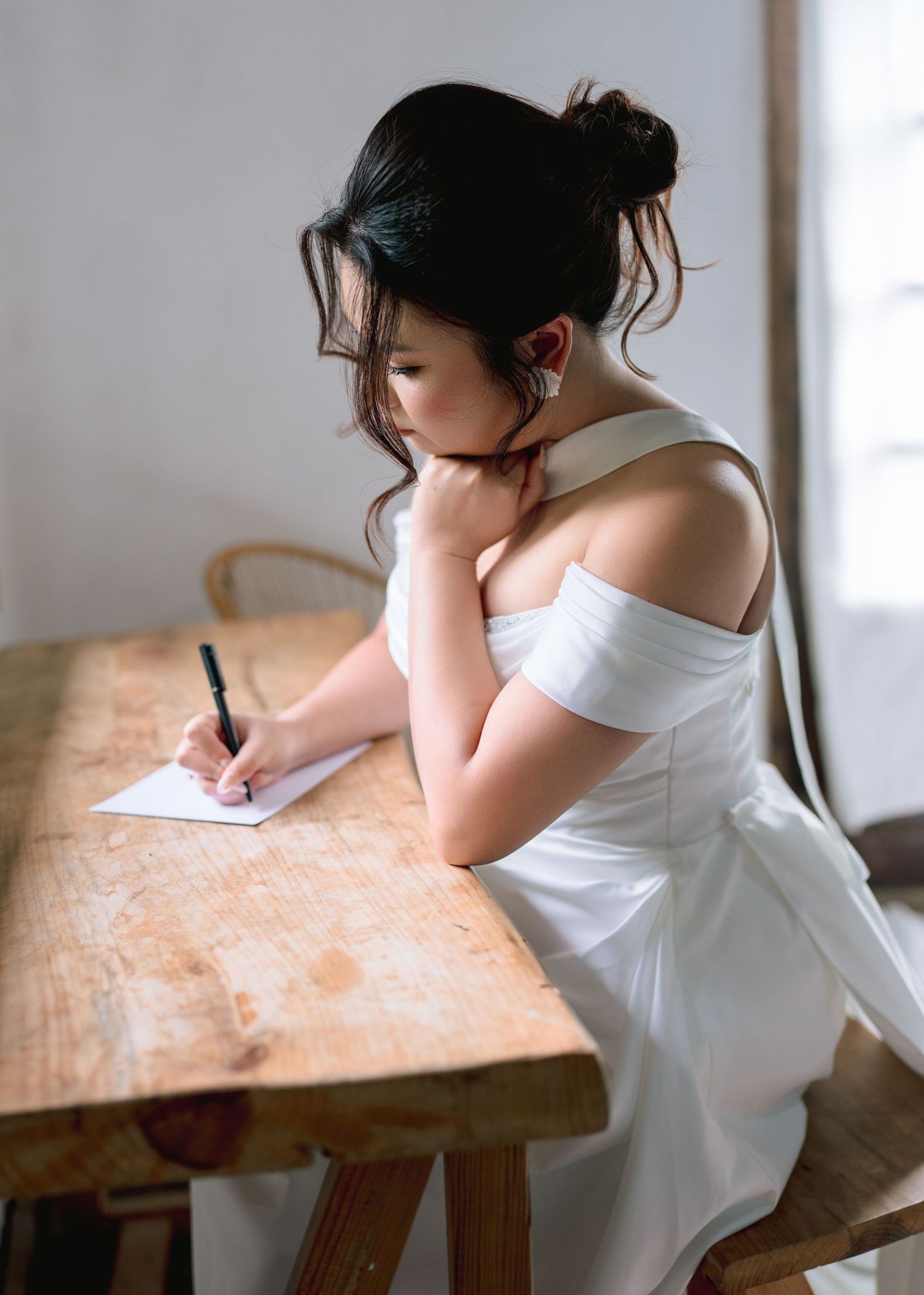 A young woman in a white off-shoulder dress sits at a wooden table, thoughtfully writing a note with a pen.