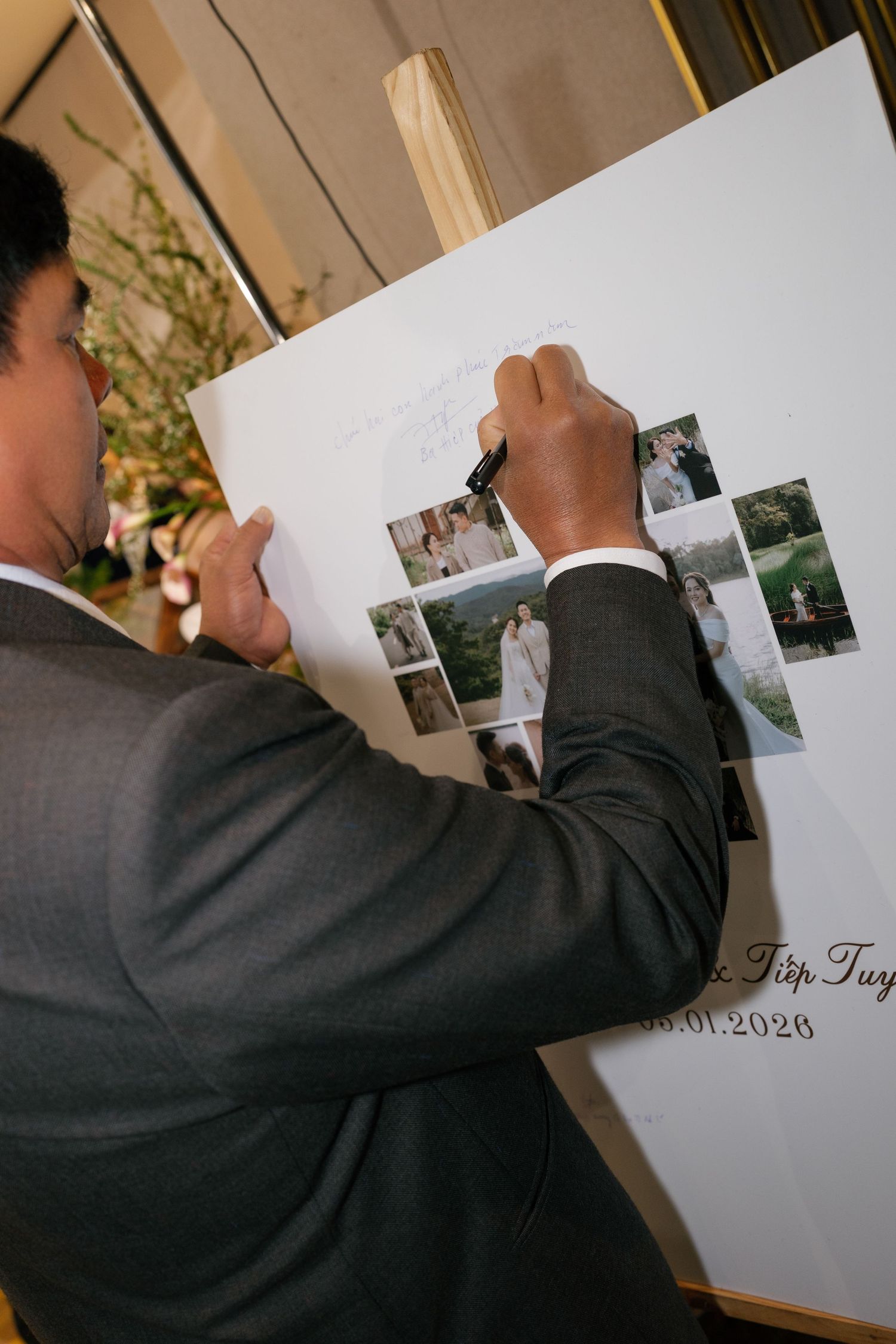 A wedding guest in a gray suit signs a photo guest book on an easel at an elegant wedding reception.