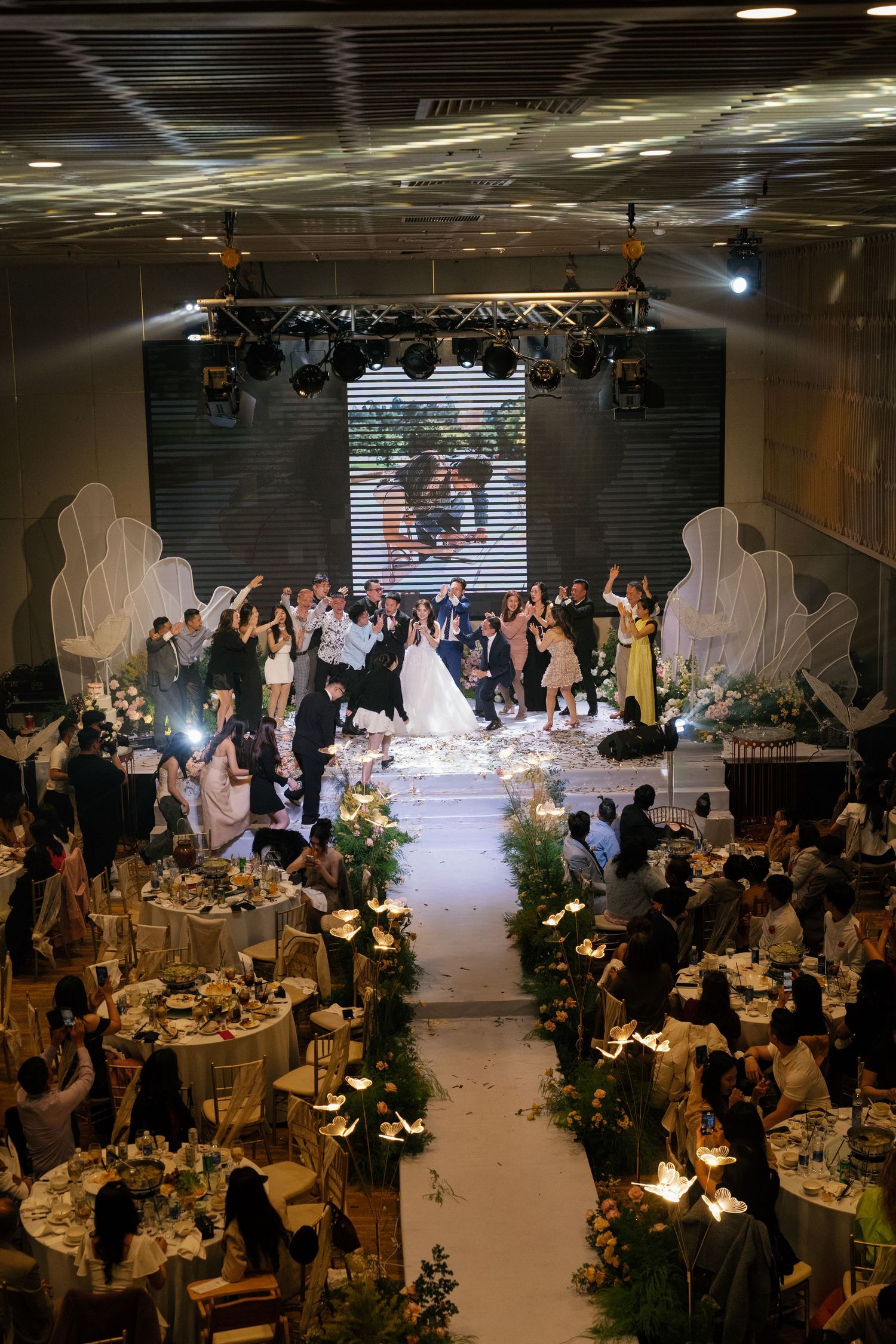 Elegant wedding reception hall with bride and groom at altar, guests celebrating at decorated tables with greenery centerpieces.