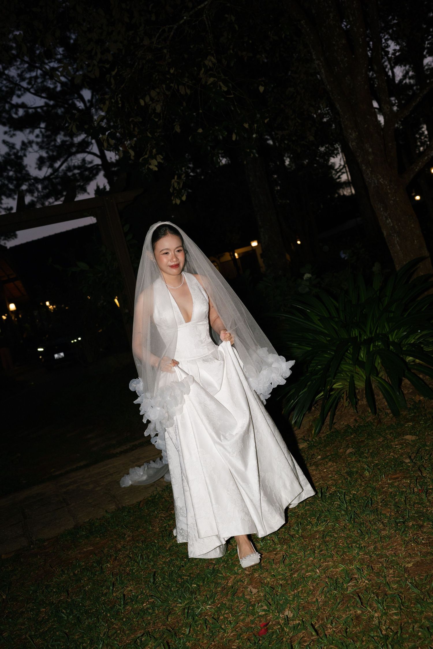 Bride in white deep-V wedding gown with ruffled veil walking through garden at night.