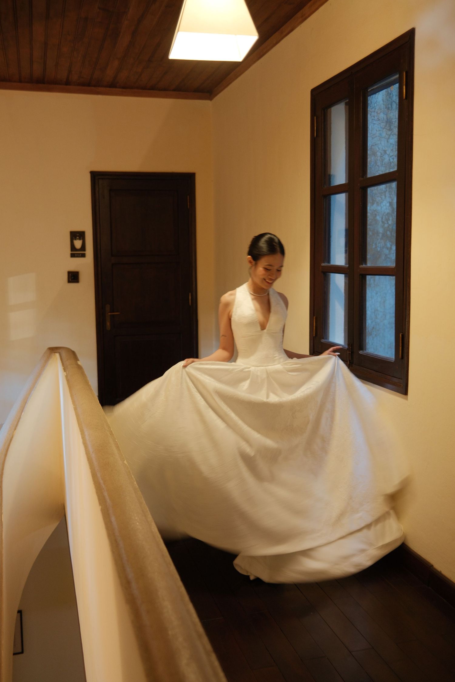 A smiling bride in a white halter wedding gown twirls on an upper-floor hallway near a window.