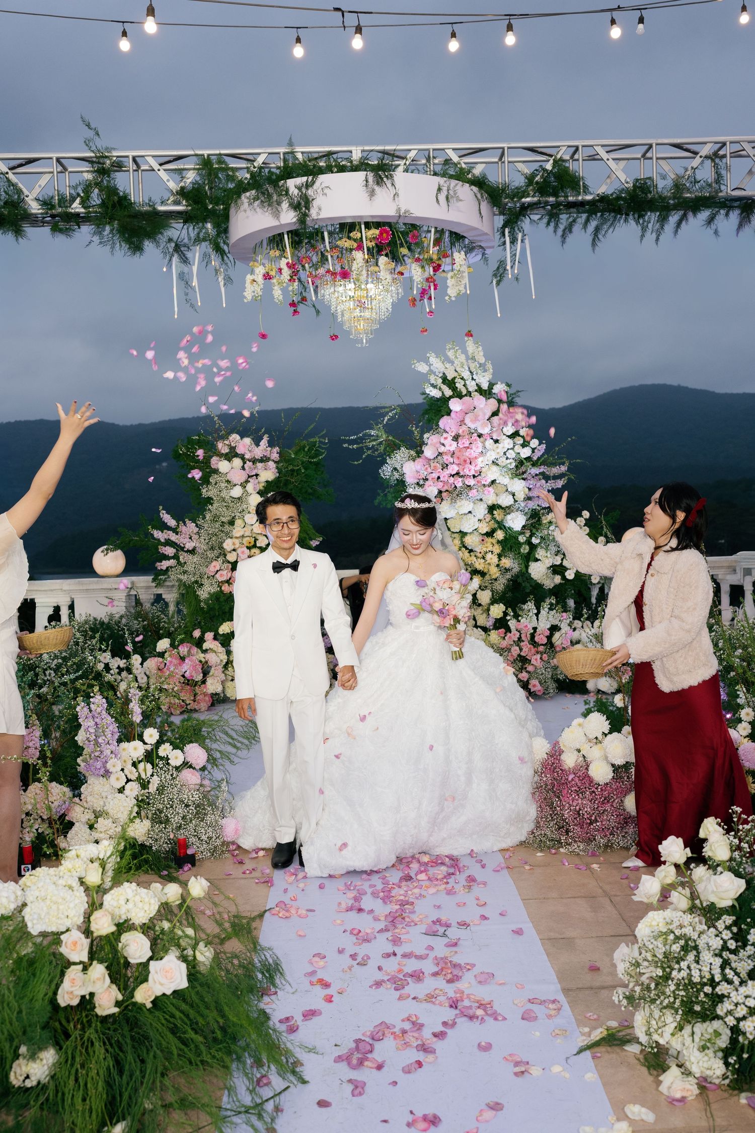 Bride and groom walk down floral aisle with pink petals thrown by guests at outdoor wedding ceremony.
